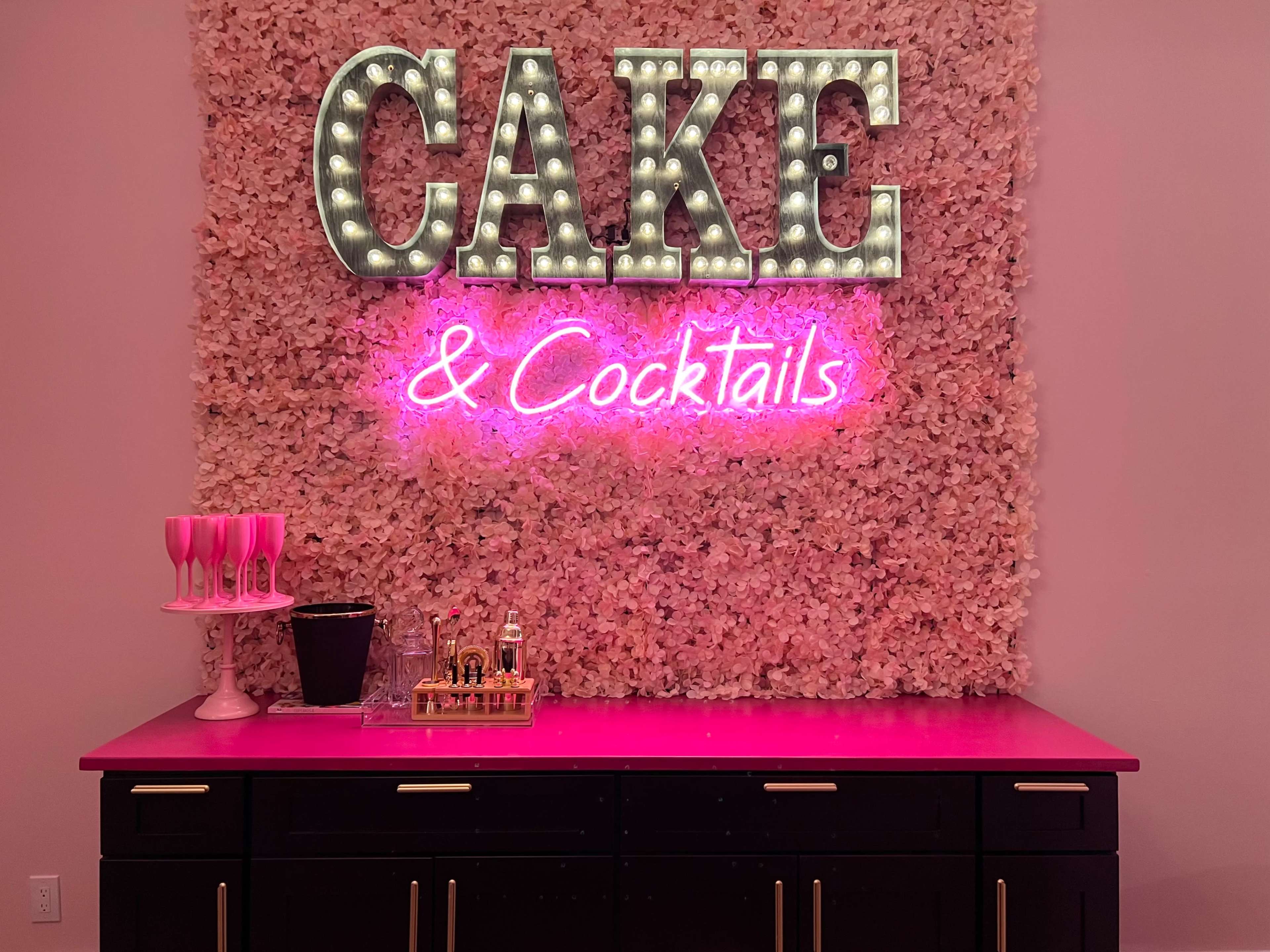 A neon sign reading "CAKE & Cocktails" is displayed on a floral backdrop above a table with champagne glasses and various drink items.