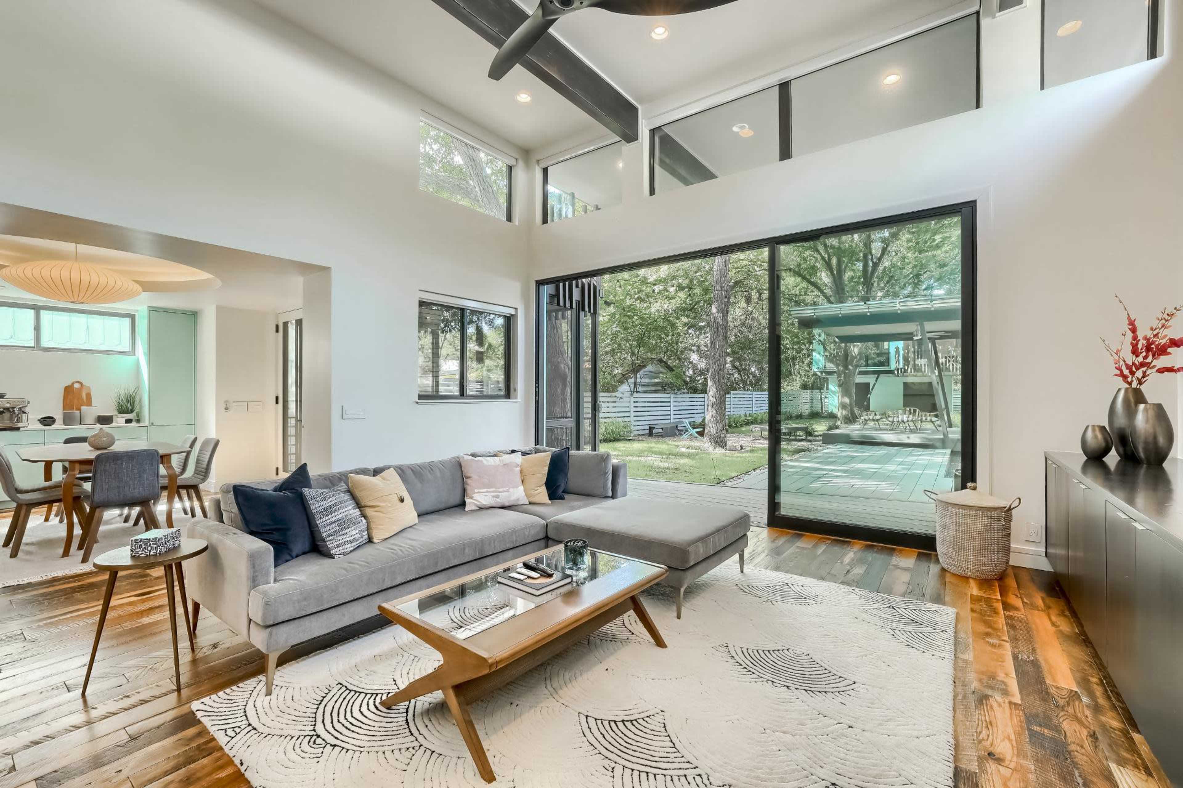 A modern living room featuring a gray sectional sofa, a wooden coffee table, and large windows overlooking a backyard with trees.