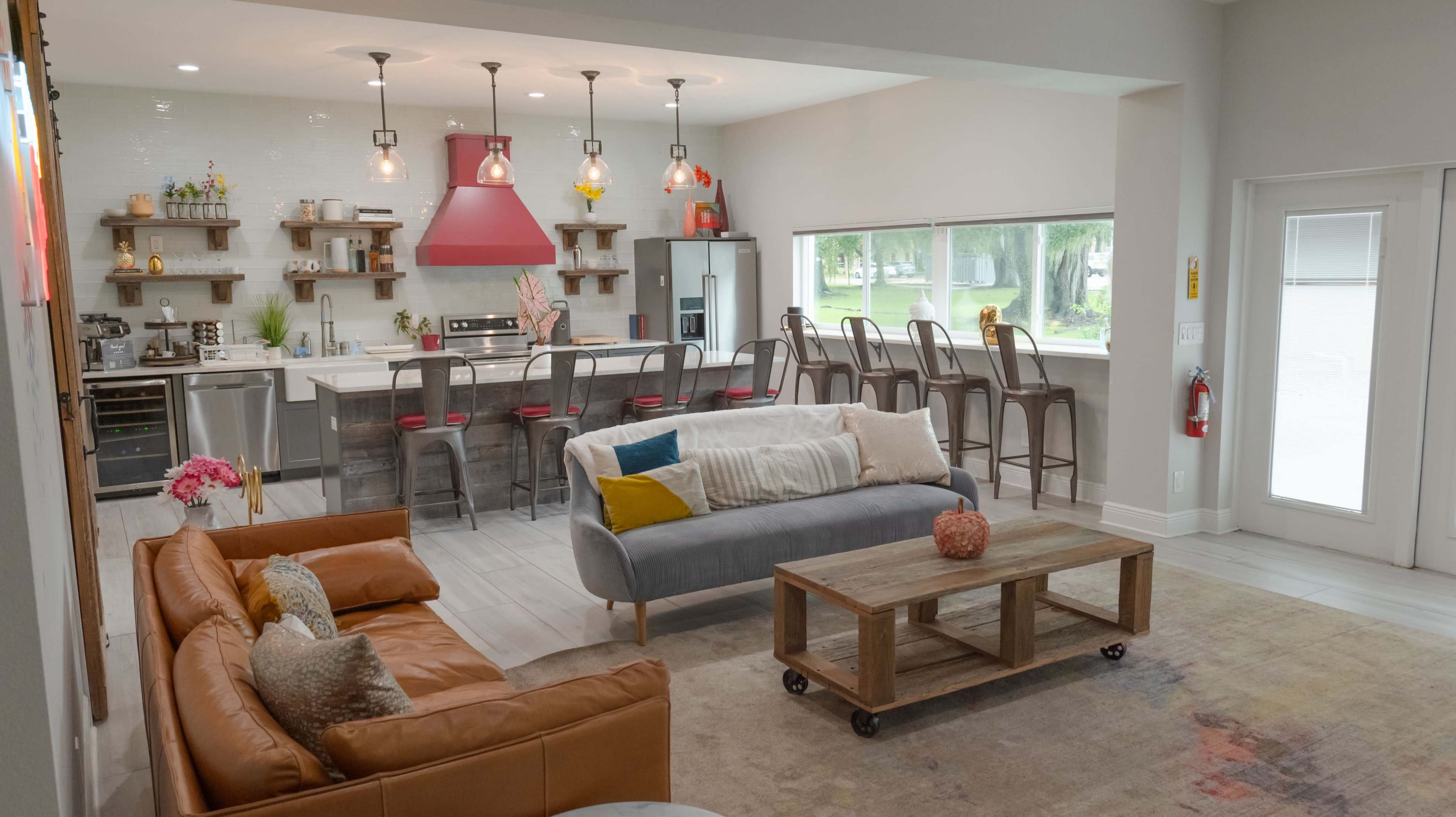 A modern, open-concept living space features a kitchen with bar seating, a cozy seating area with diverse furniture, and large windows allowing natural light.