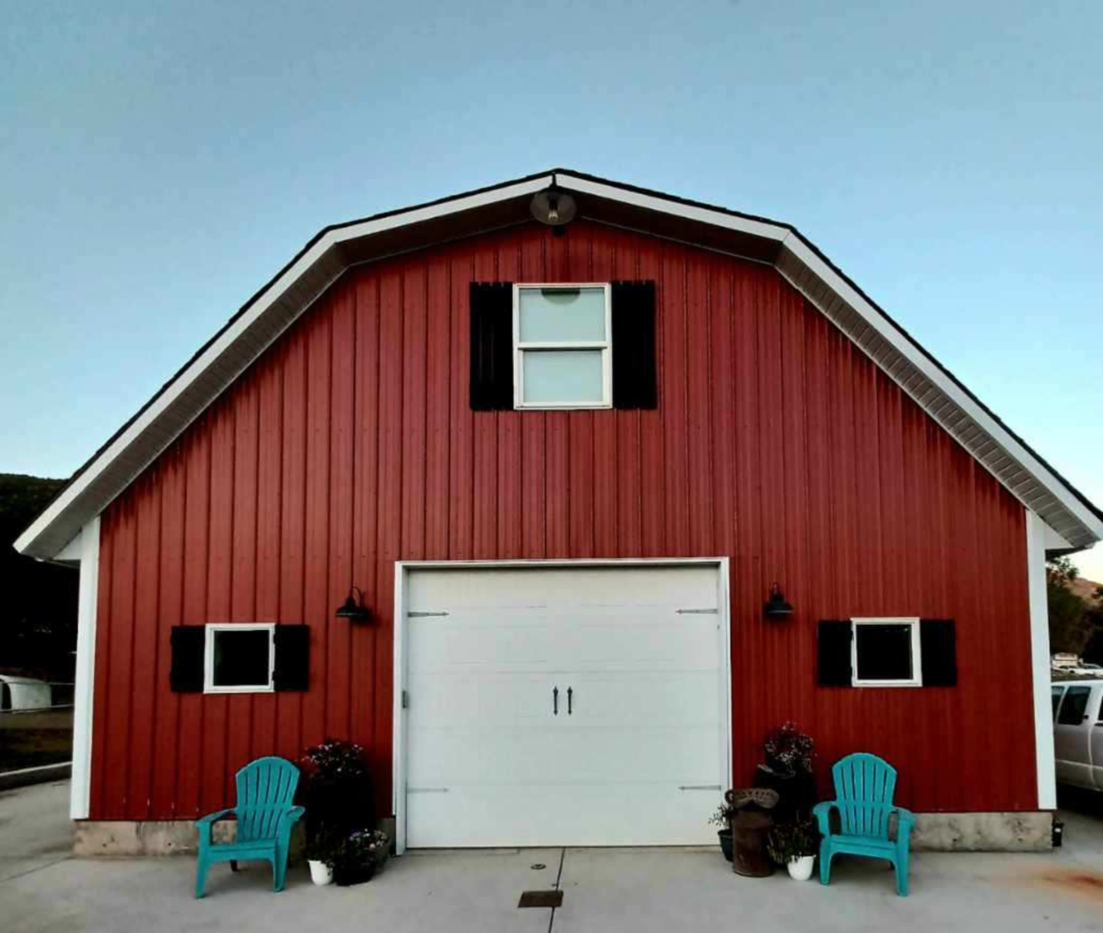 A red barn with a white garage door, flanked by black shutters and two blue chairs on either side.