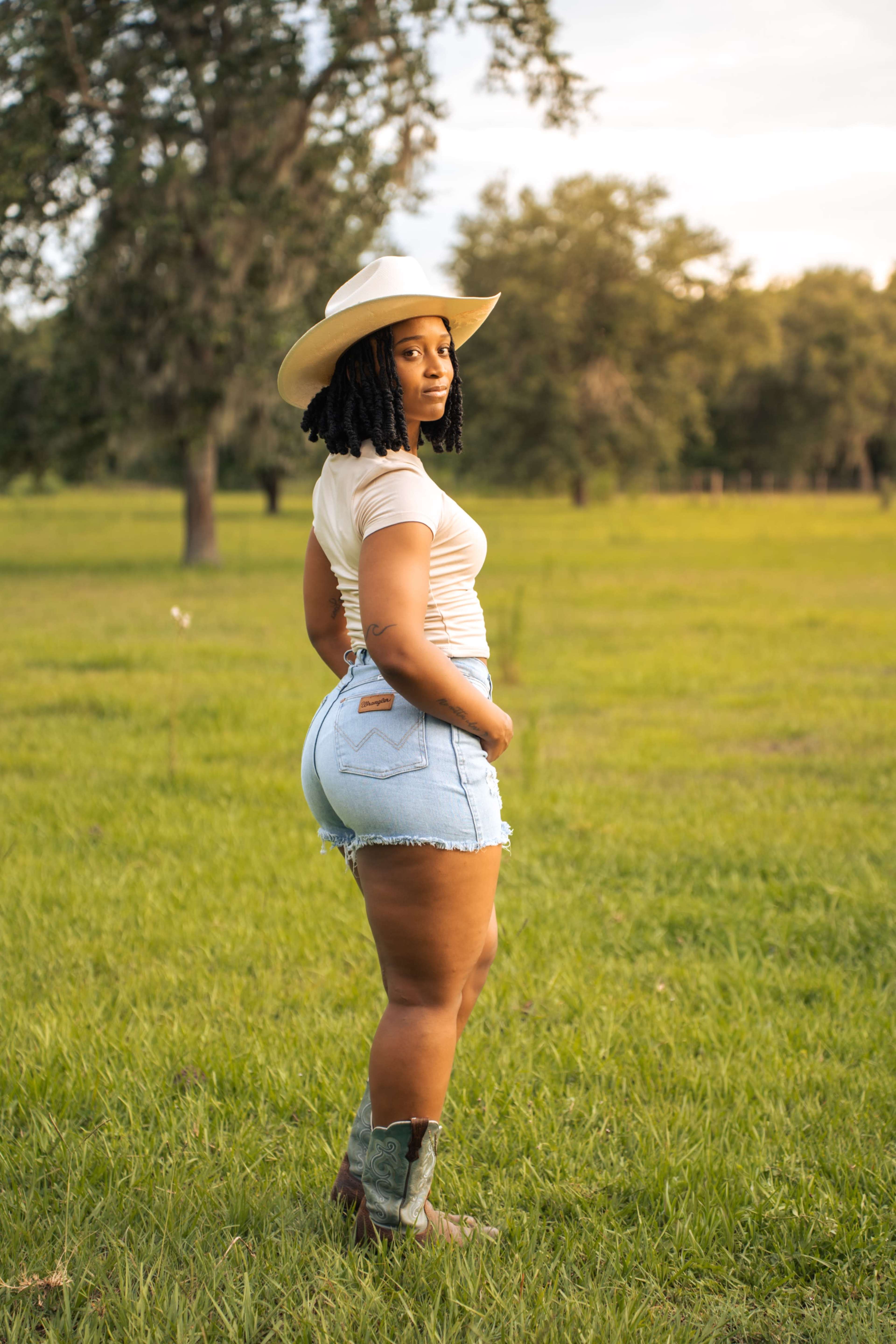 A woman wearing a white top, denim shorts, and cowboy boots stands in a grassy field, facing away and looking back over her shoulder.