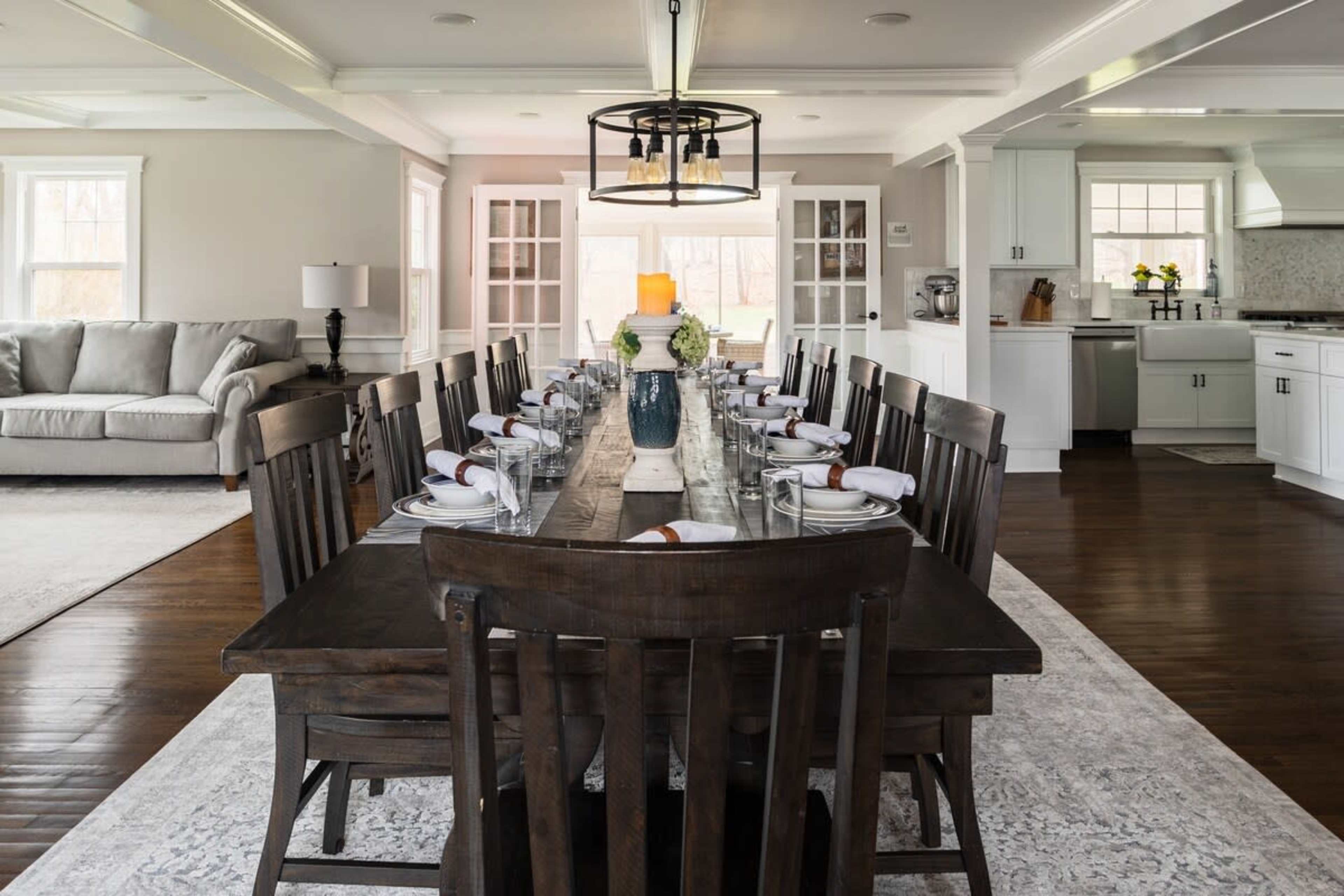 A long wooden dining table is set with glasses and napkins in a bright open kitchen and dining area featuring large windows and a comfortable seating area.