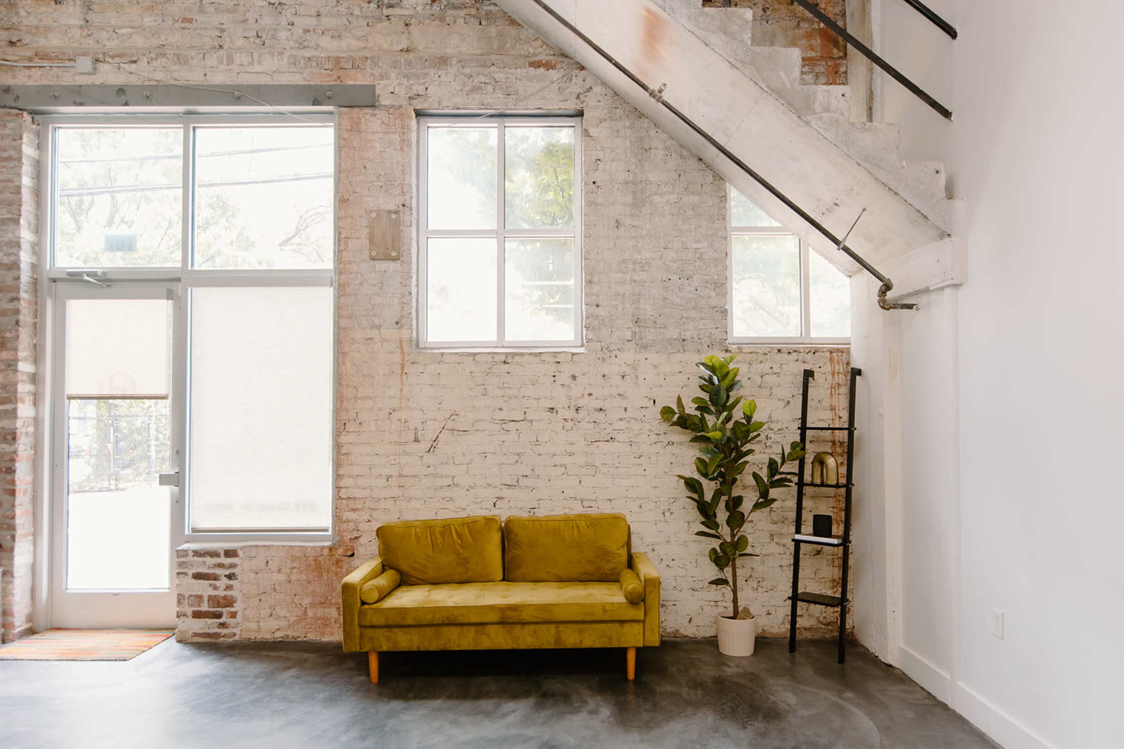 A modern interior space with a yellow sofa, a potted plant, and a stairway against a brick and concrete wall.