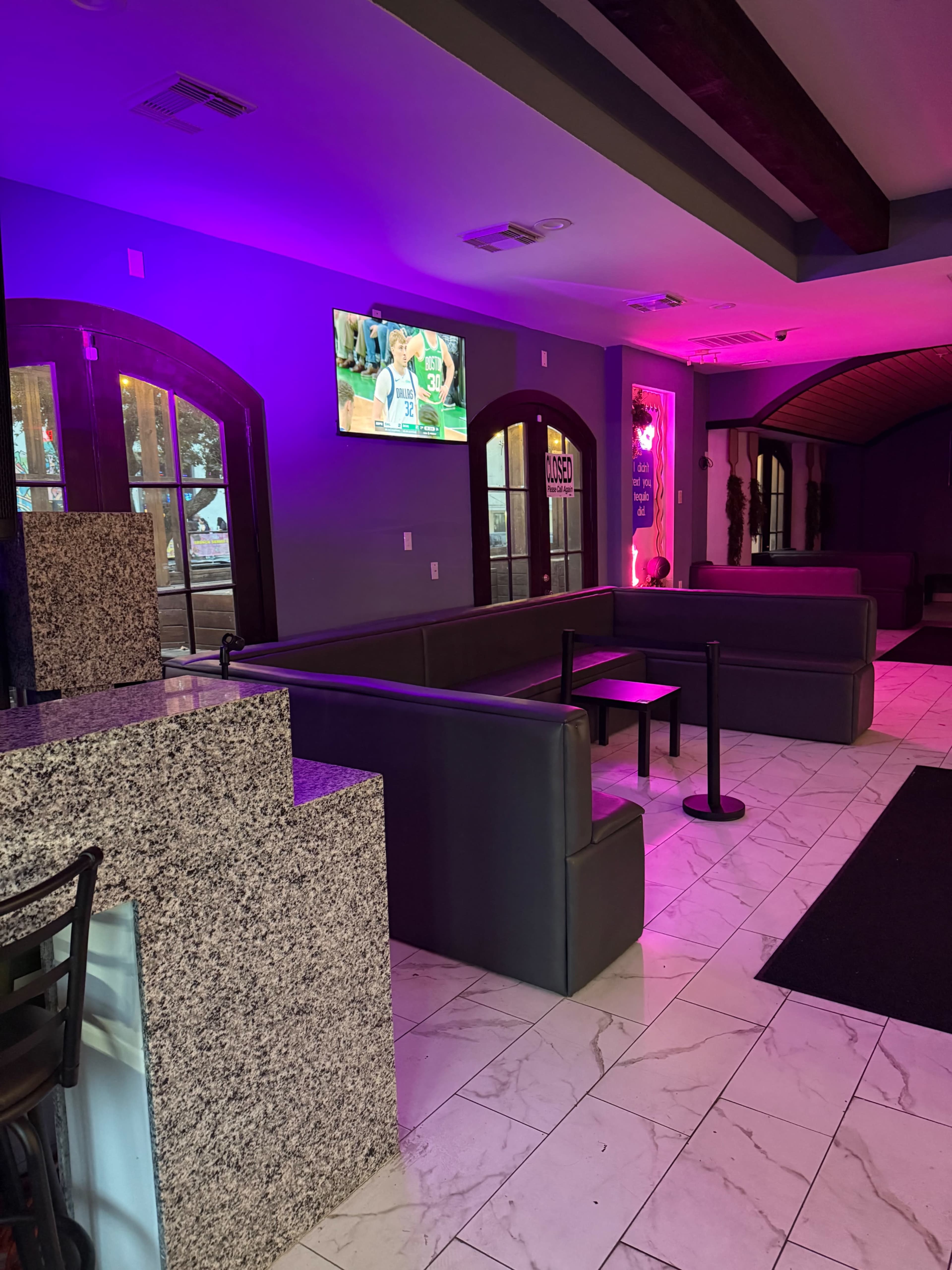 The image shows a modern lounge area with dark sofas, a small table, and a television displaying a sports game, illuminated by purple lighting.