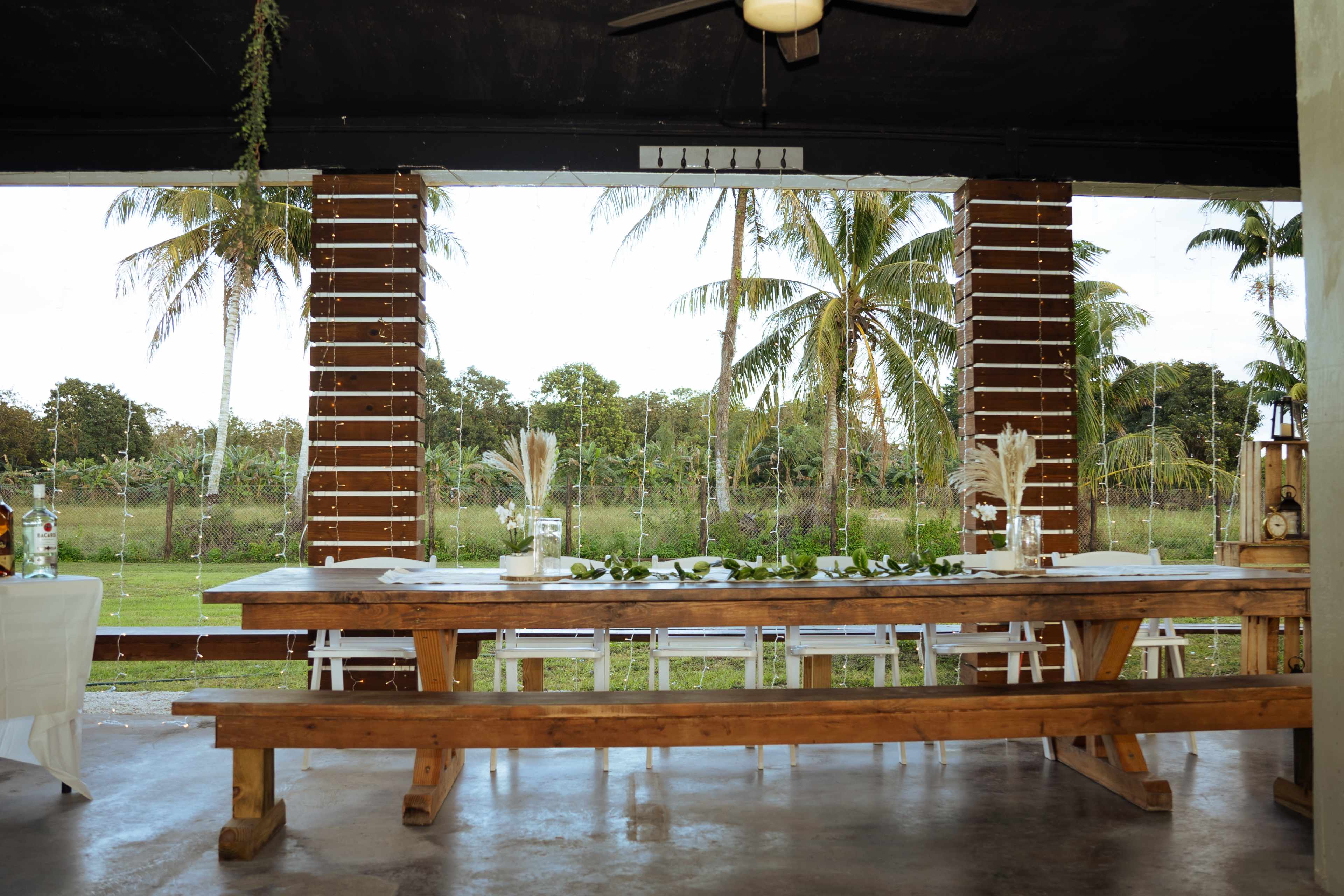 A long wooden dining table is set up under a covered porch with palm trees and a field visible in the background.