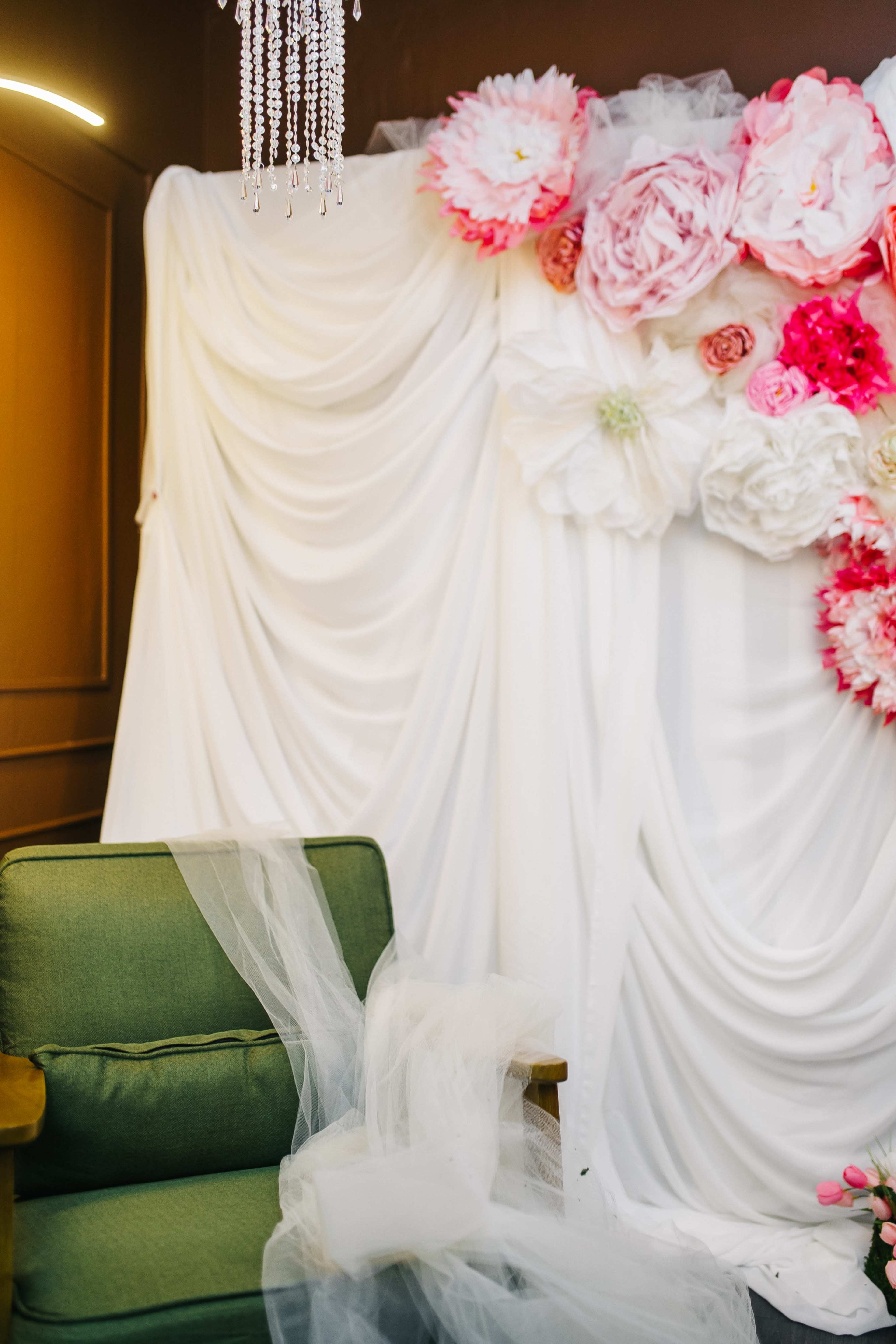 The image shows a decorated backdrop of white fabric adorned with large, colorful paper flowers, beside a green upholstered chair.