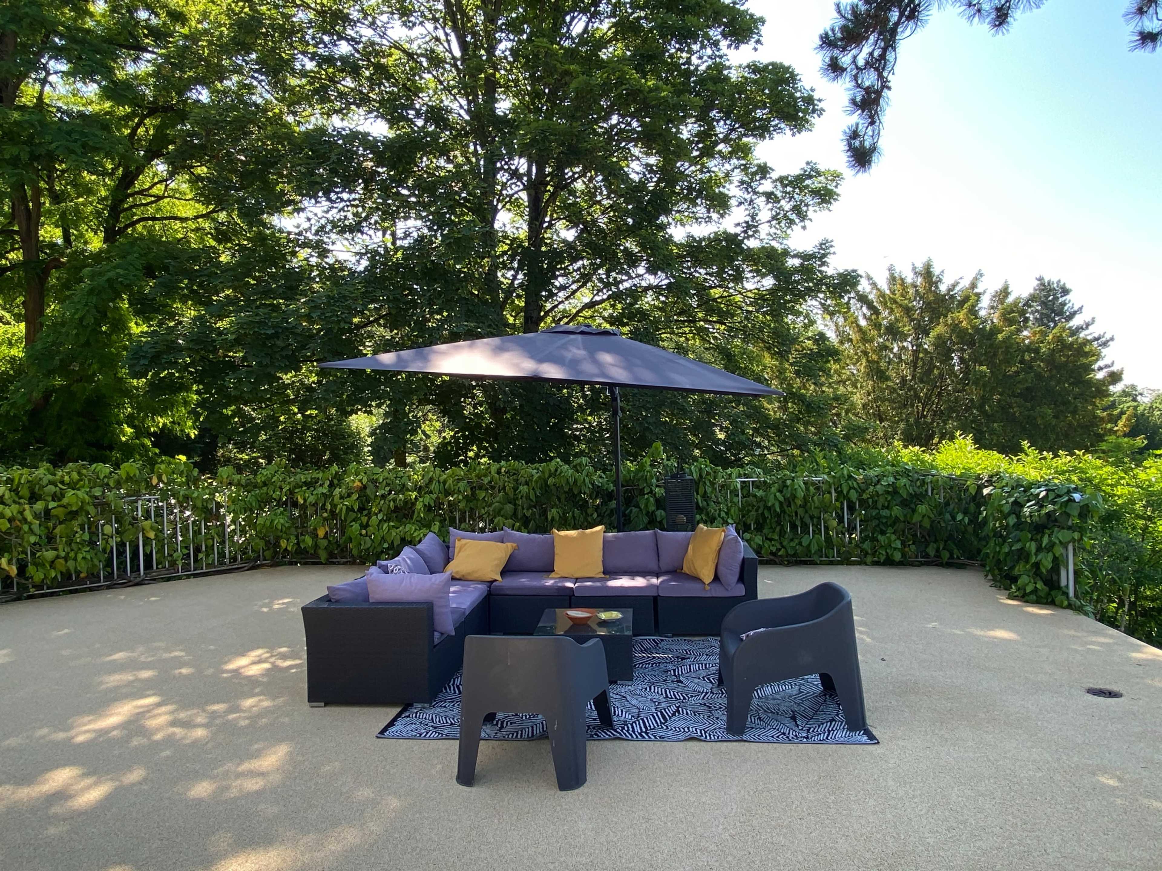 A patio area is furnished with a sectional sofa, two chairs, a coffee table, and a large umbrella, surrounded by greenery.
