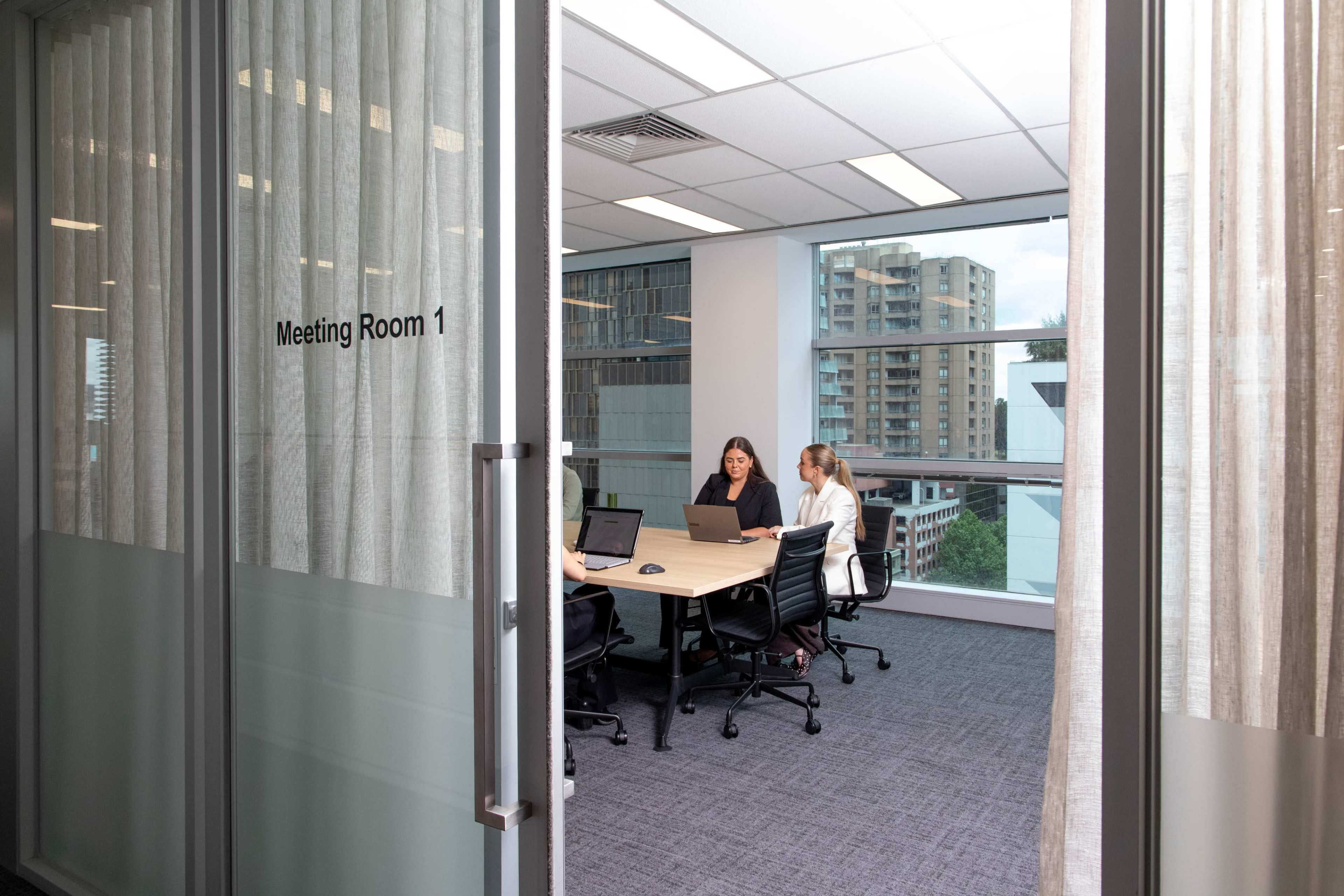 A group of three people is engaged in a discussion around a table in a glass-walled meeting room.