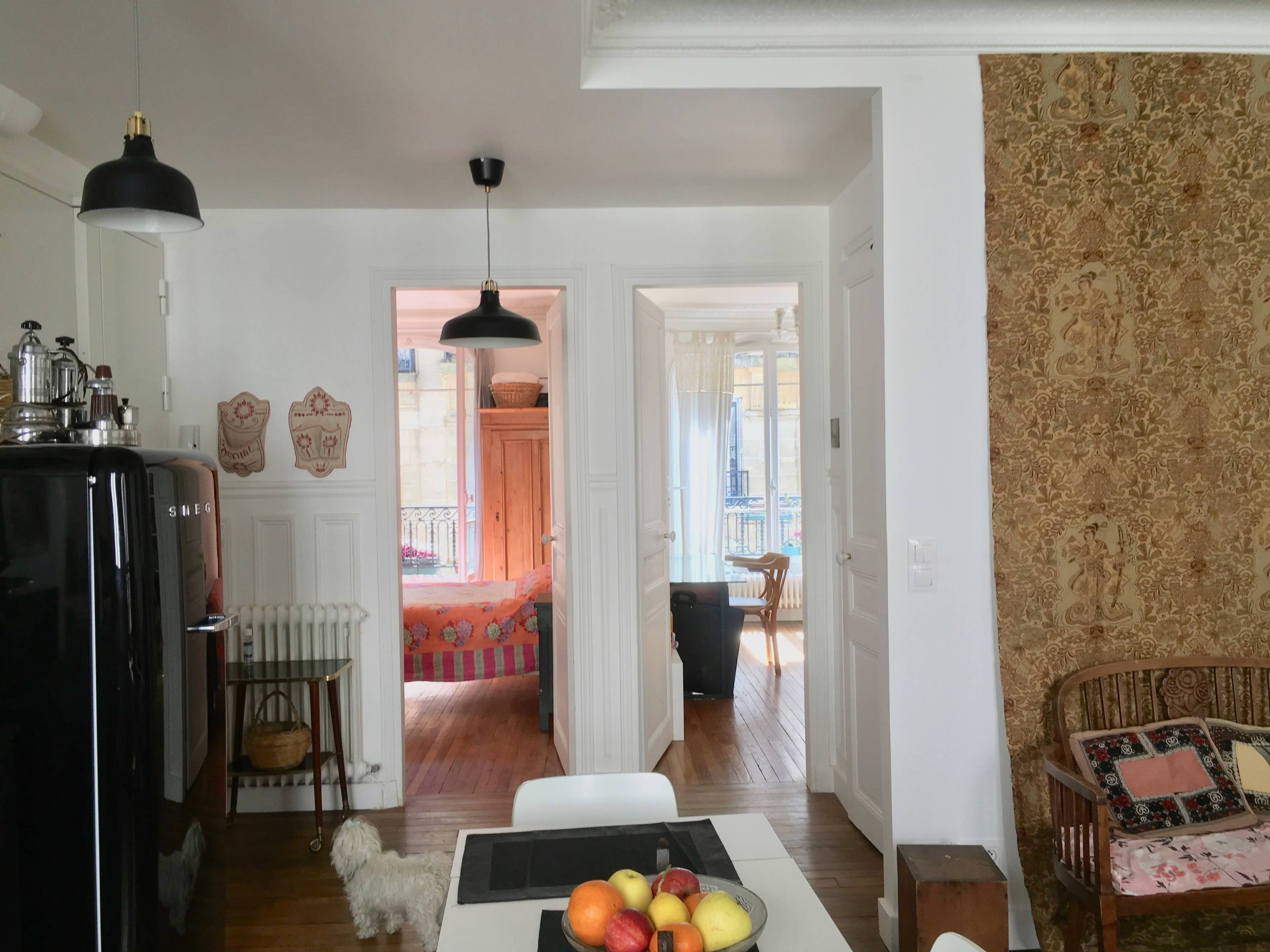 A bright kitchen with a table featuring a bowl of fruit, leading to a hallway that connects to another room with a visible bed.