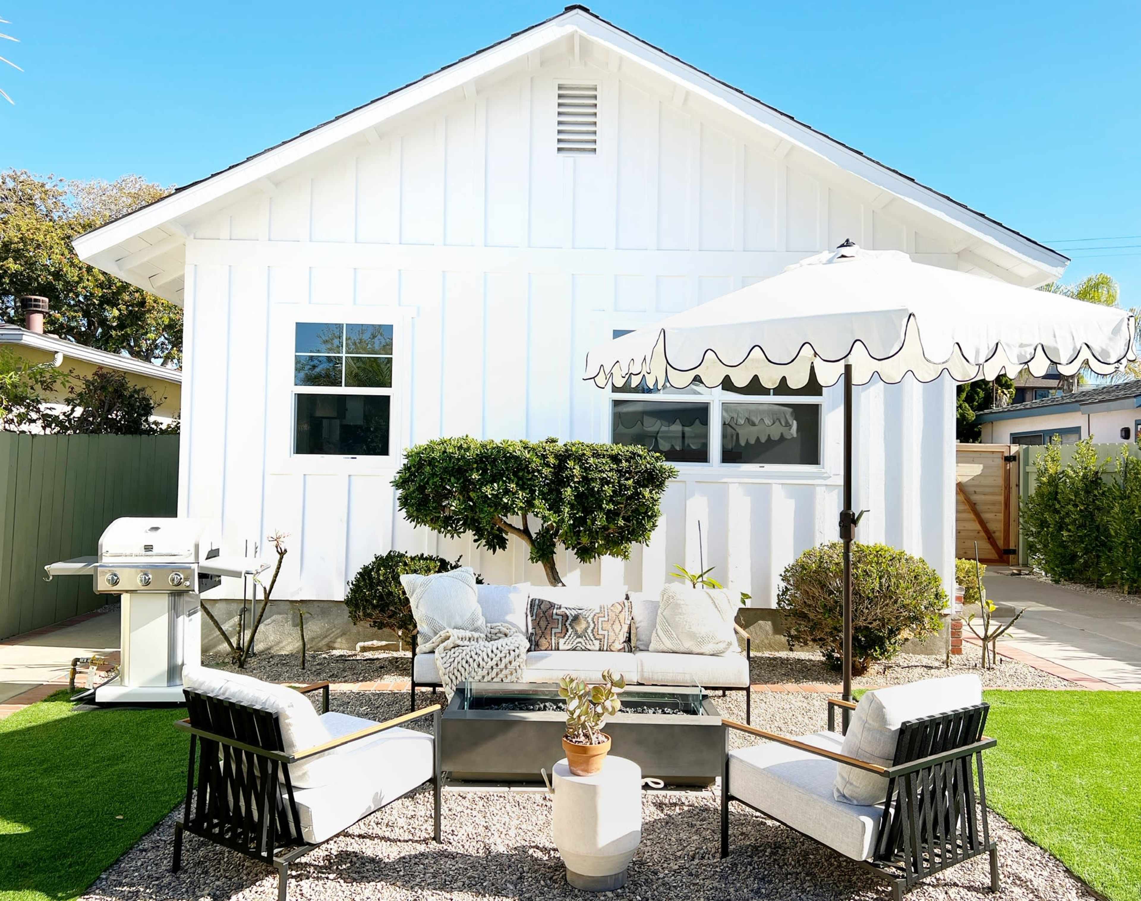 A backyard patio setup with a grill, seating area, and an umbrella near a white house.