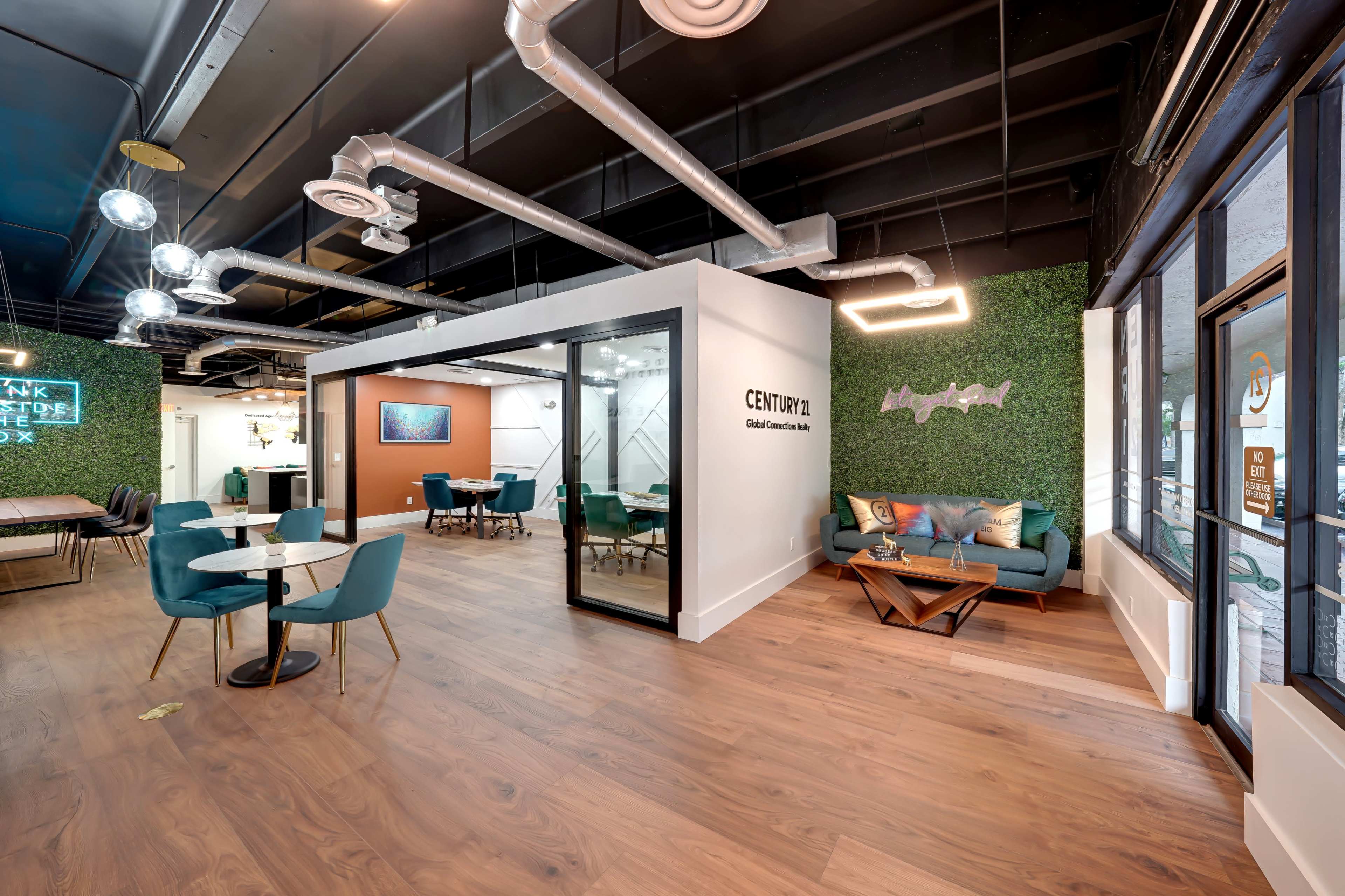 A modern office space featuring a communal area with green wall accents, tables, chairs, and glass-walled meeting rooms.