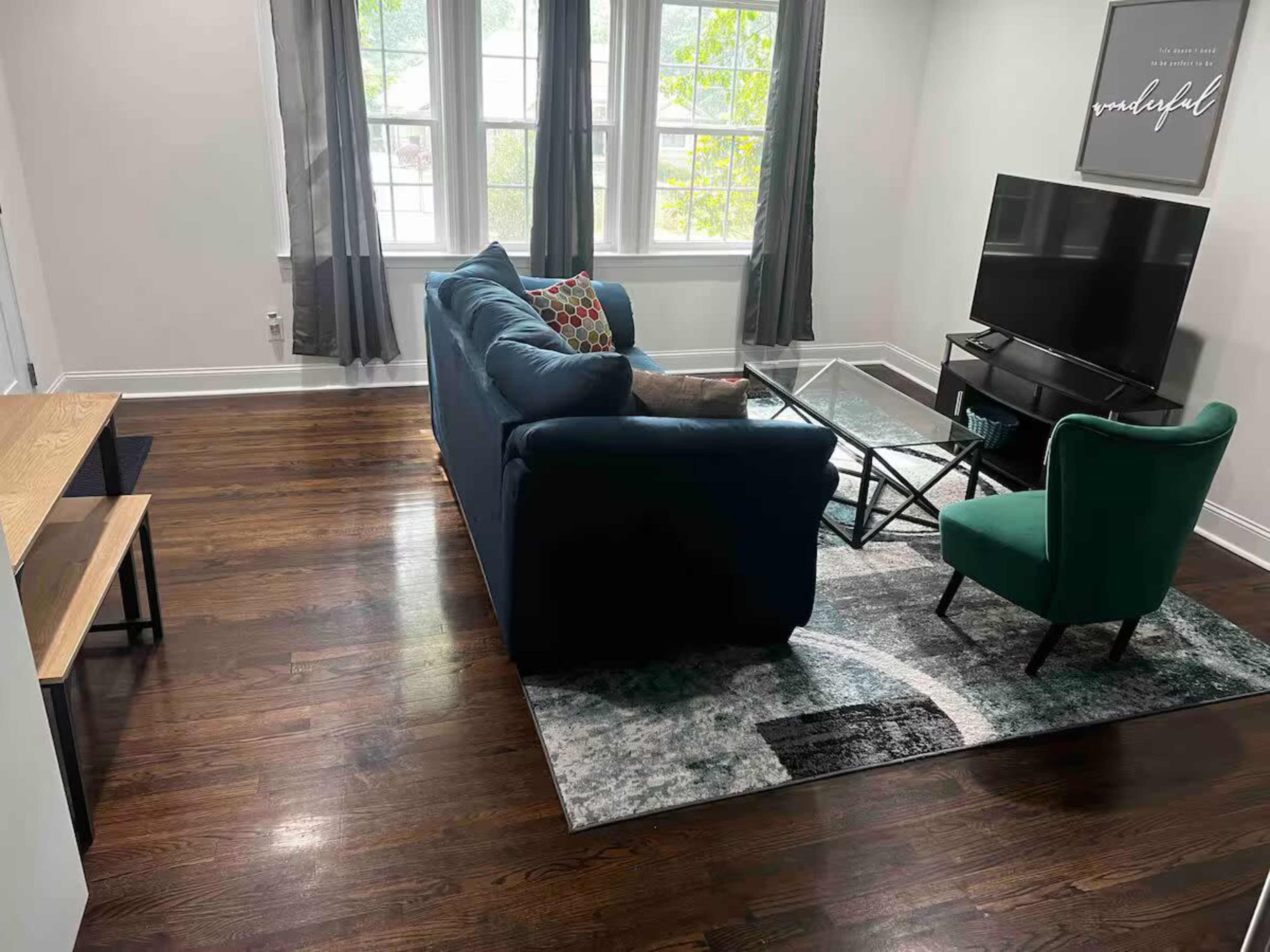 A living room features a blue sofa, a green chair, a glass coffee table, a television, and large windows allowing natural light to enter.