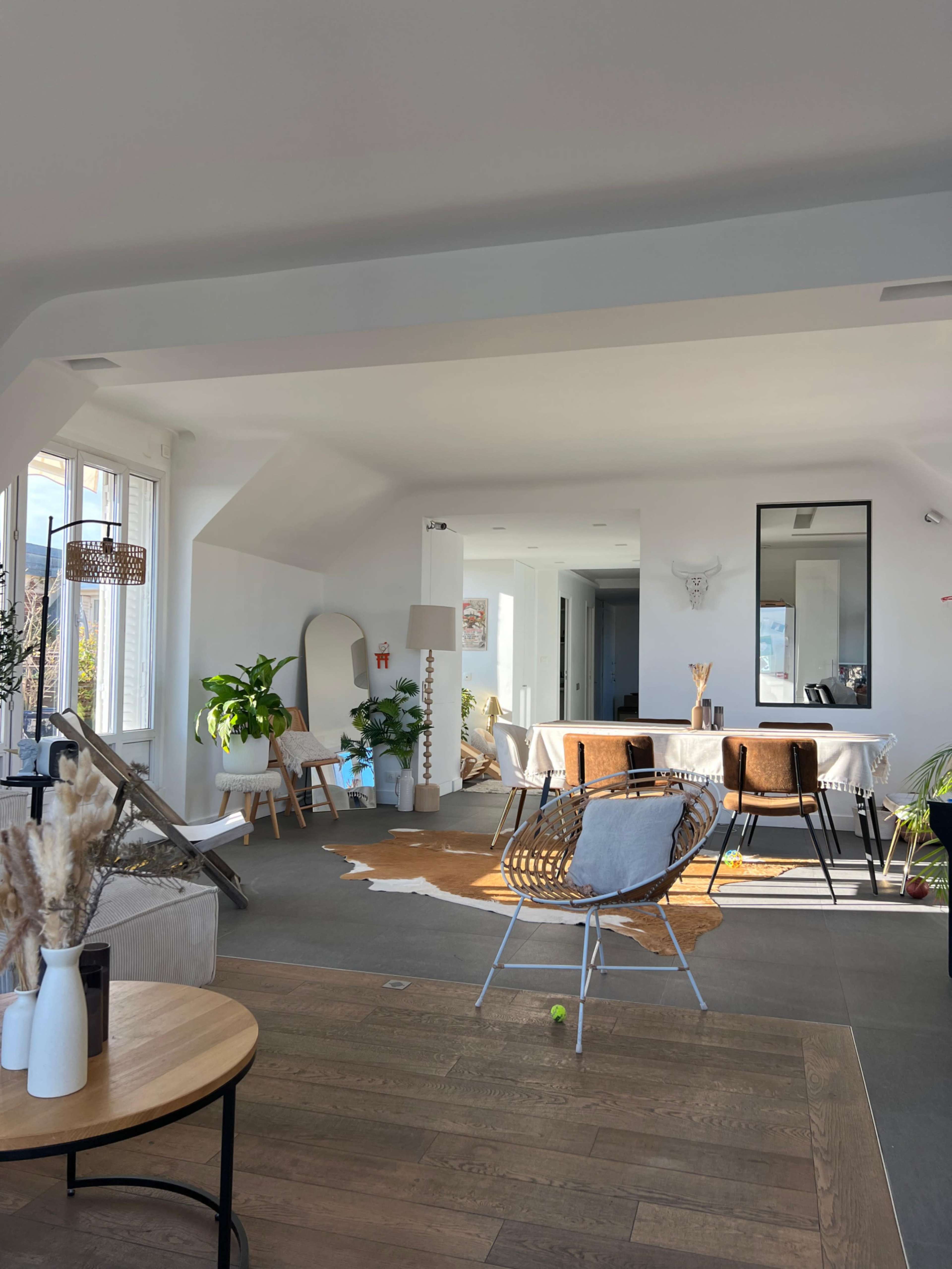 The image shows a bright, modern living space featuring a dining area with a table and chairs, a lounge area with seating, and large windows allowing natural light to enter.