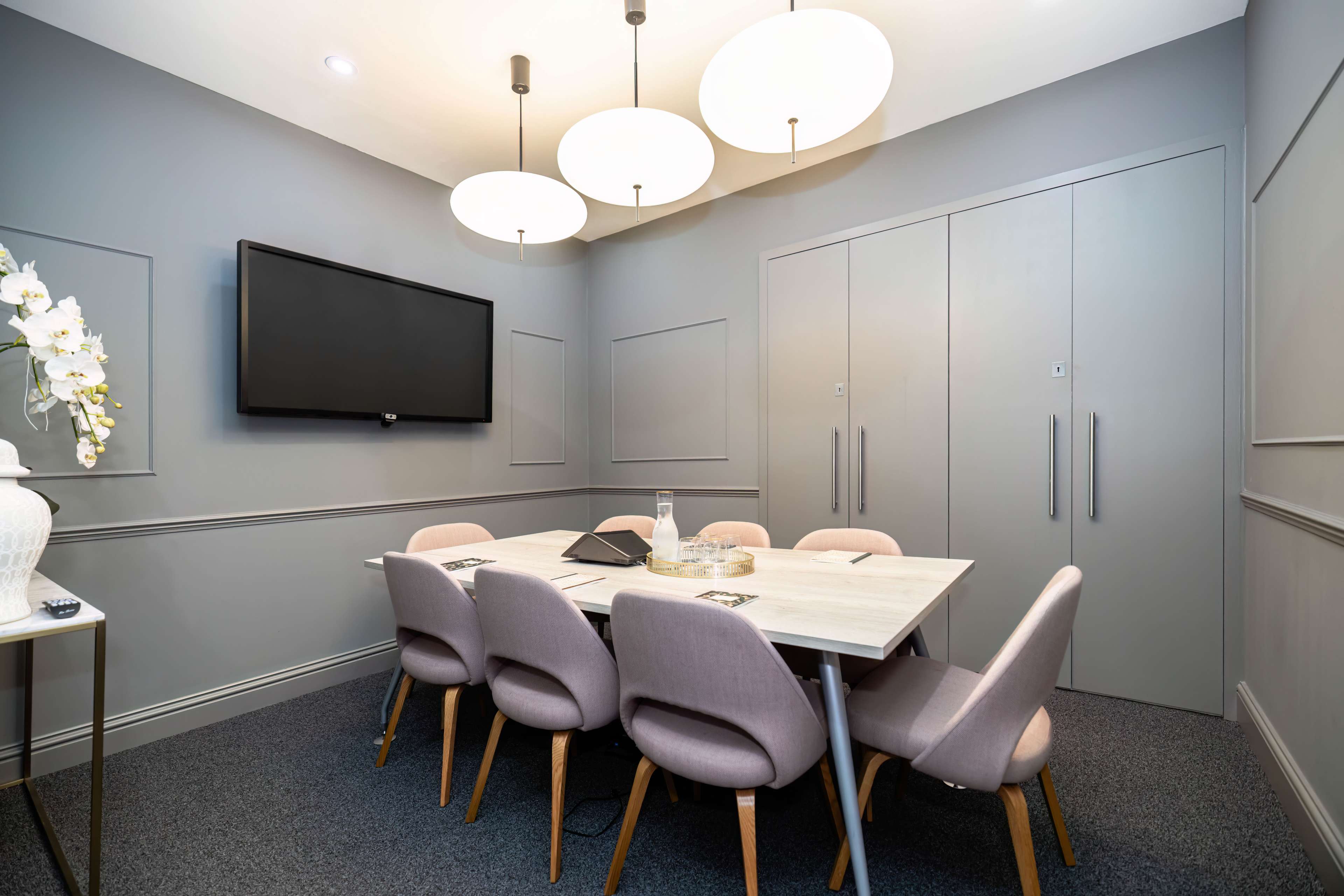 A modern meeting room features a table surrounded by six upholstered chairs, a wall-mounted TV, and large pendant lights overhead.