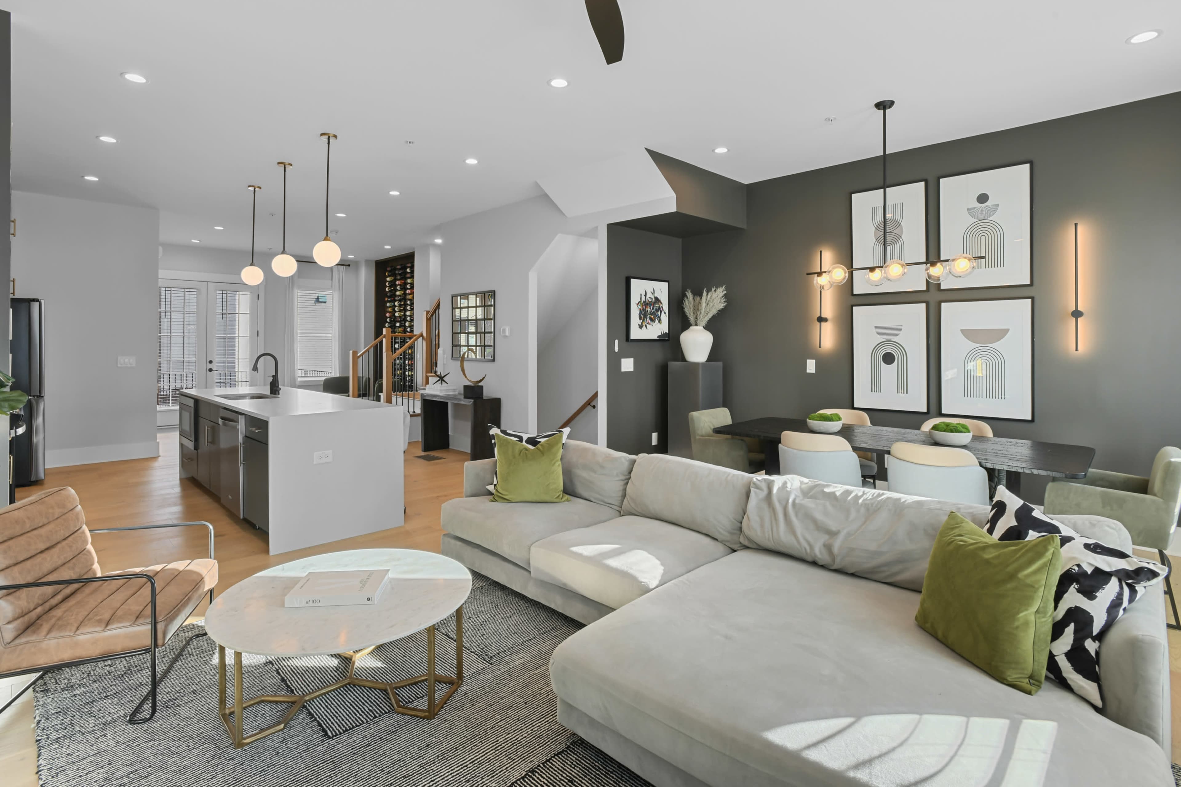 The image shows a modern living room with a light-colored sectional sofa, a circular coffee table, and a dining area featuring a dark table and decorative wall art.