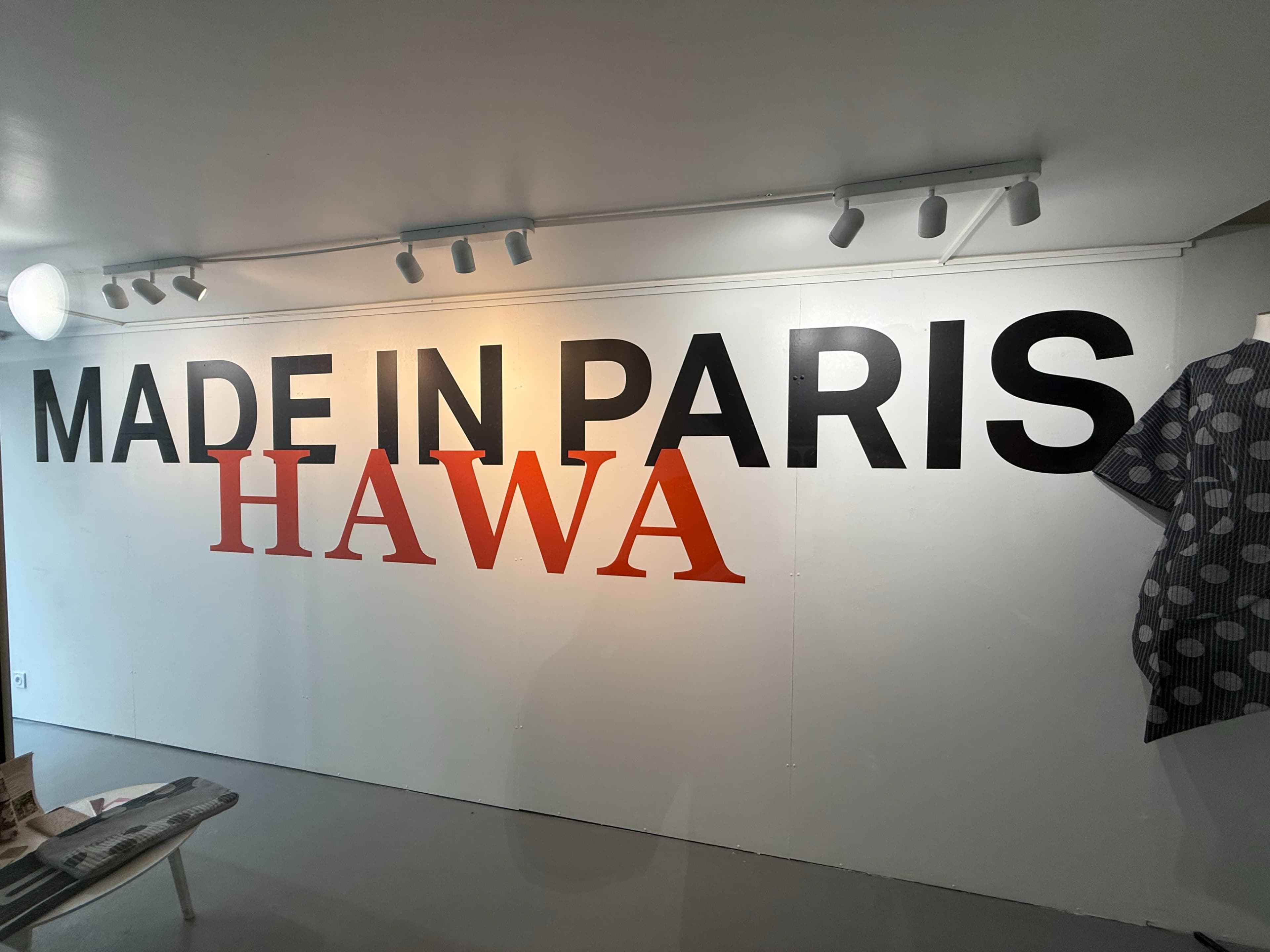 The image shows a wall with the text "MADE IN PARIS" in large black letters and "HAWA" in bold red letters, alongside a patterned garment displayed on the right.