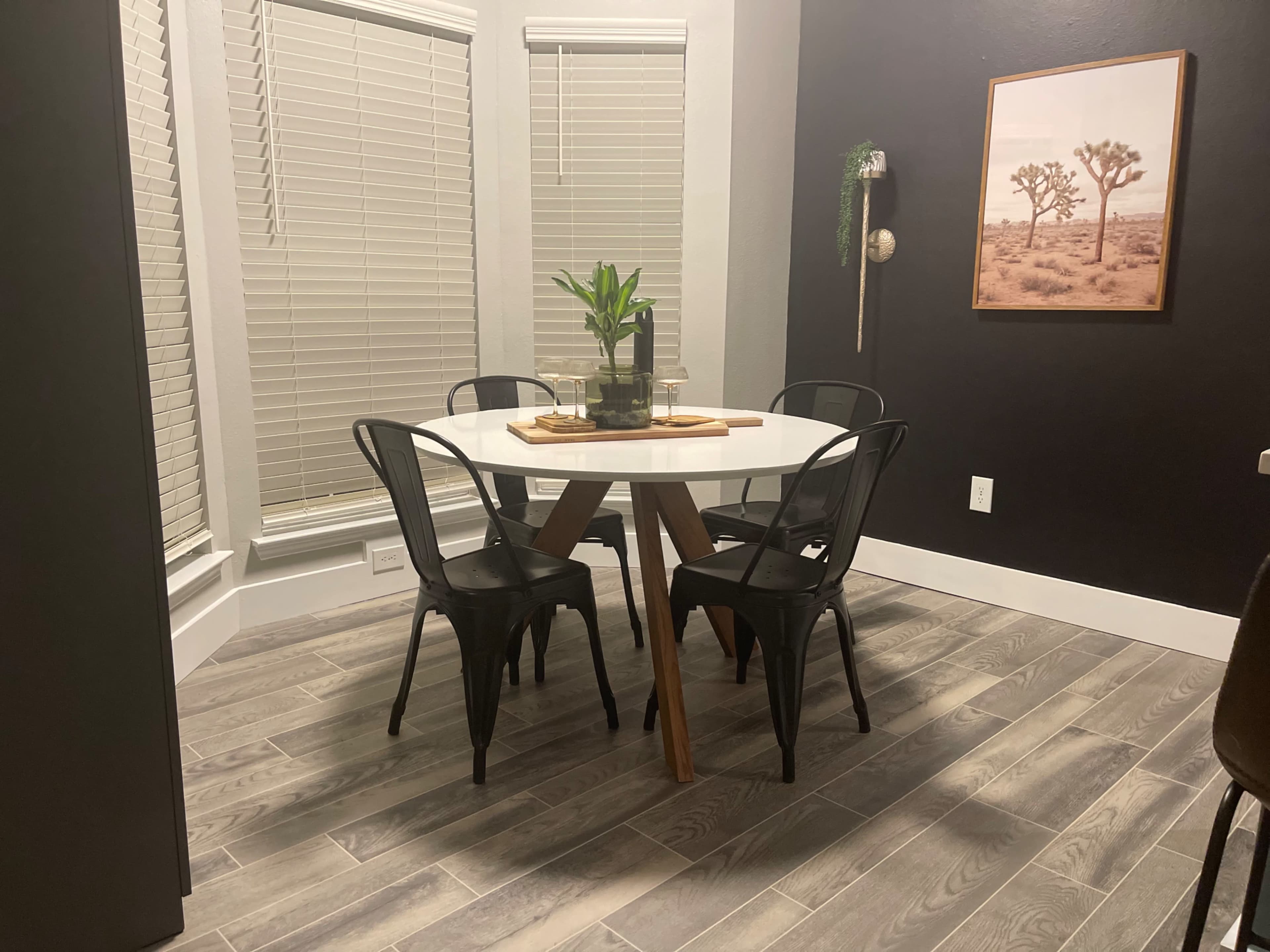 A round white table with a wooden base is surrounded by four black chairs in a room with gray walls and large windows.