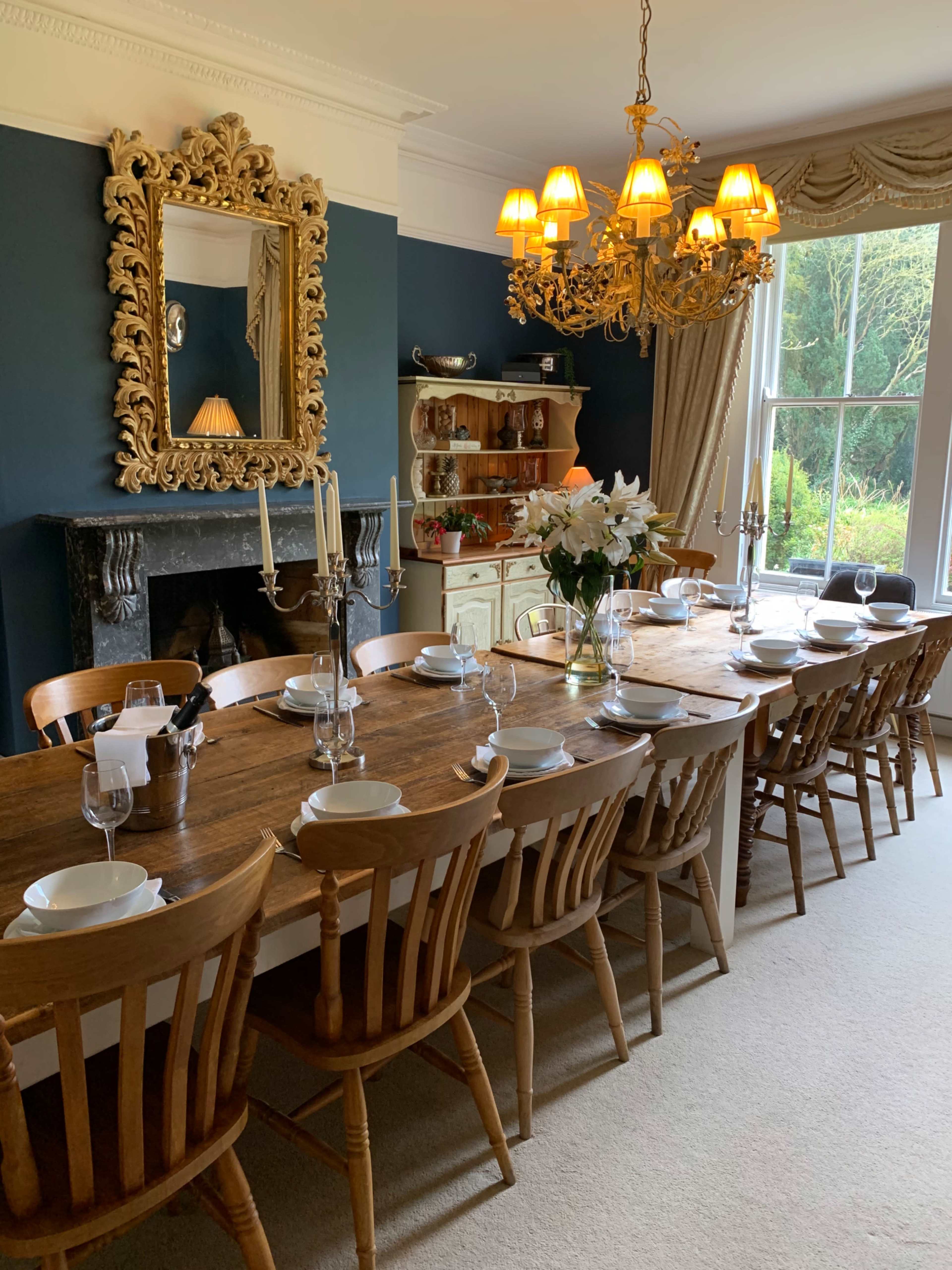 A long wooden dining table is set with plates and glasses, surrounded by wooden chairs in a room featuring a large ornate mirror and a chandelier.