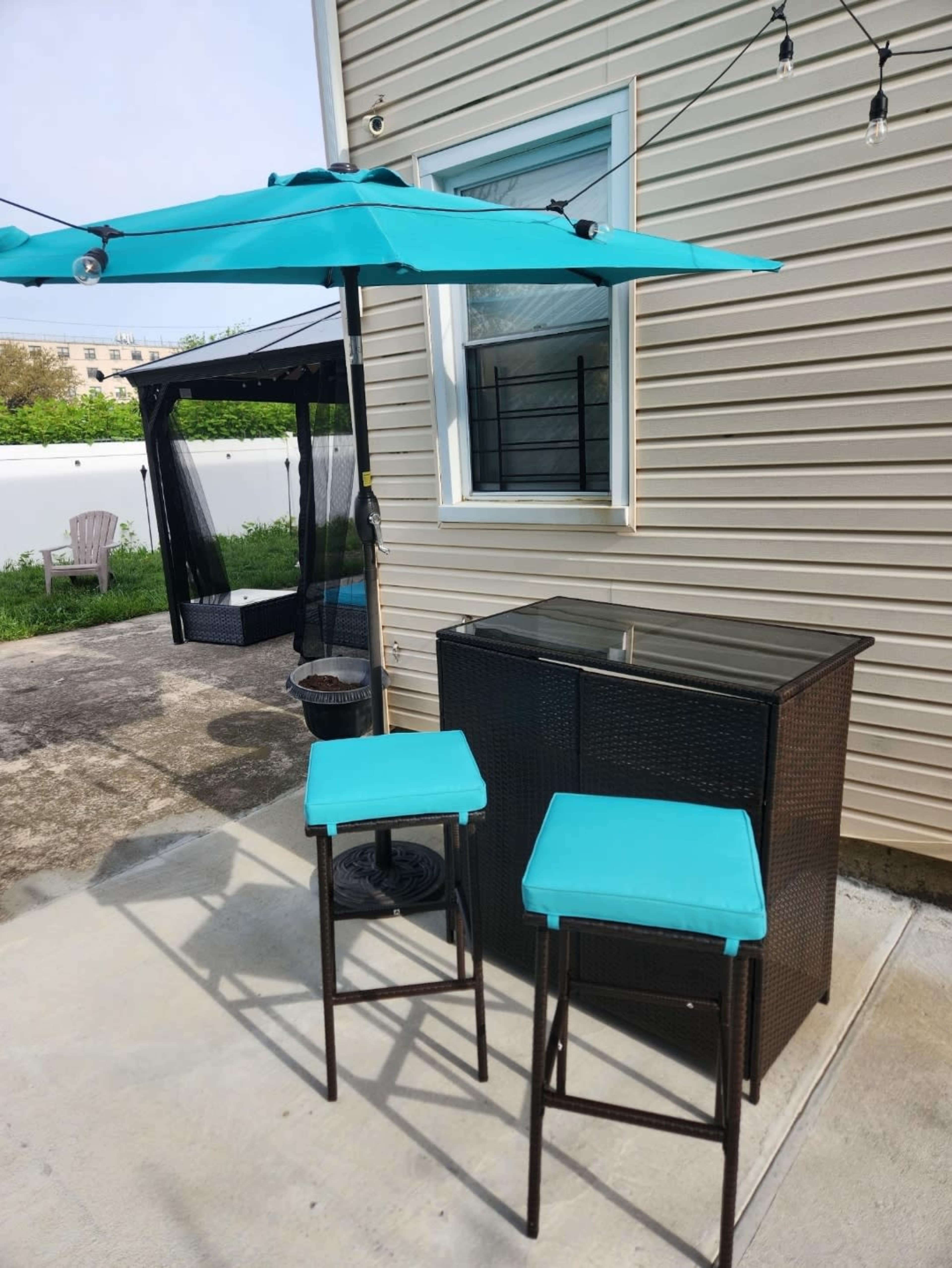A patio area features a small bar table with two teal stools and a blue umbrella, set against a beige siding backdrop.