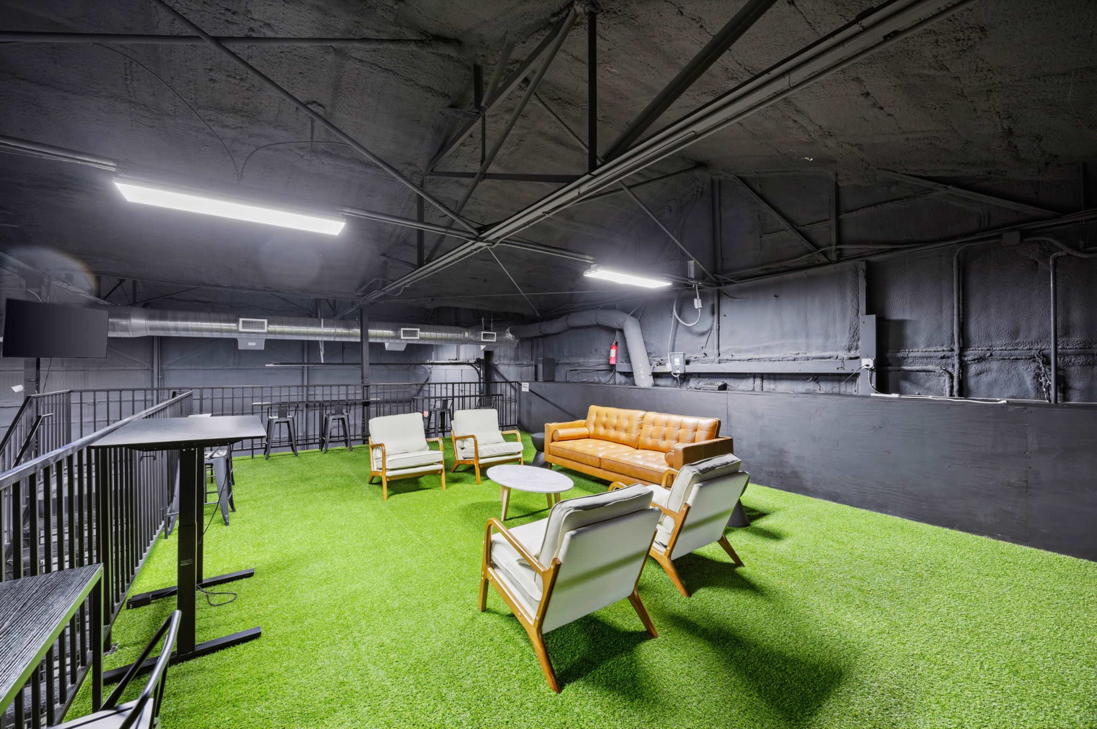 The image shows a modern lounge area featuring a tan leather sofa and white chairs on artificial grass flooring beneath a high ceiling with exposed beams and visible lighting.