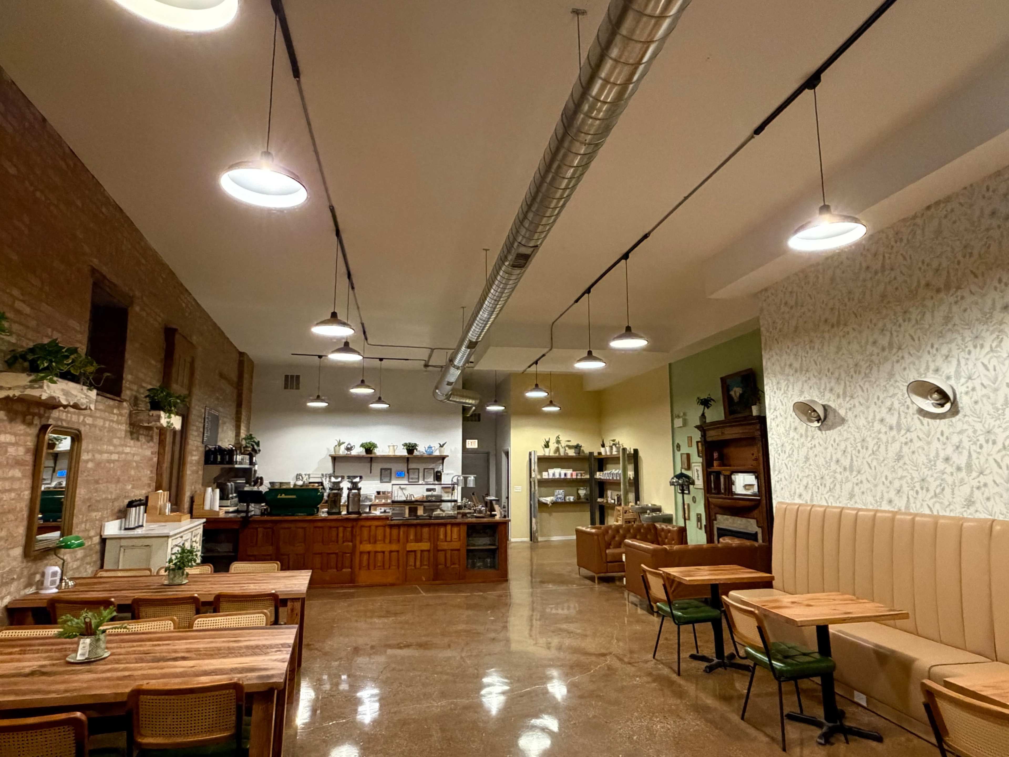 The image shows a spacious café interior with wooden tables, an open counter area, and warm, pendant lighting.