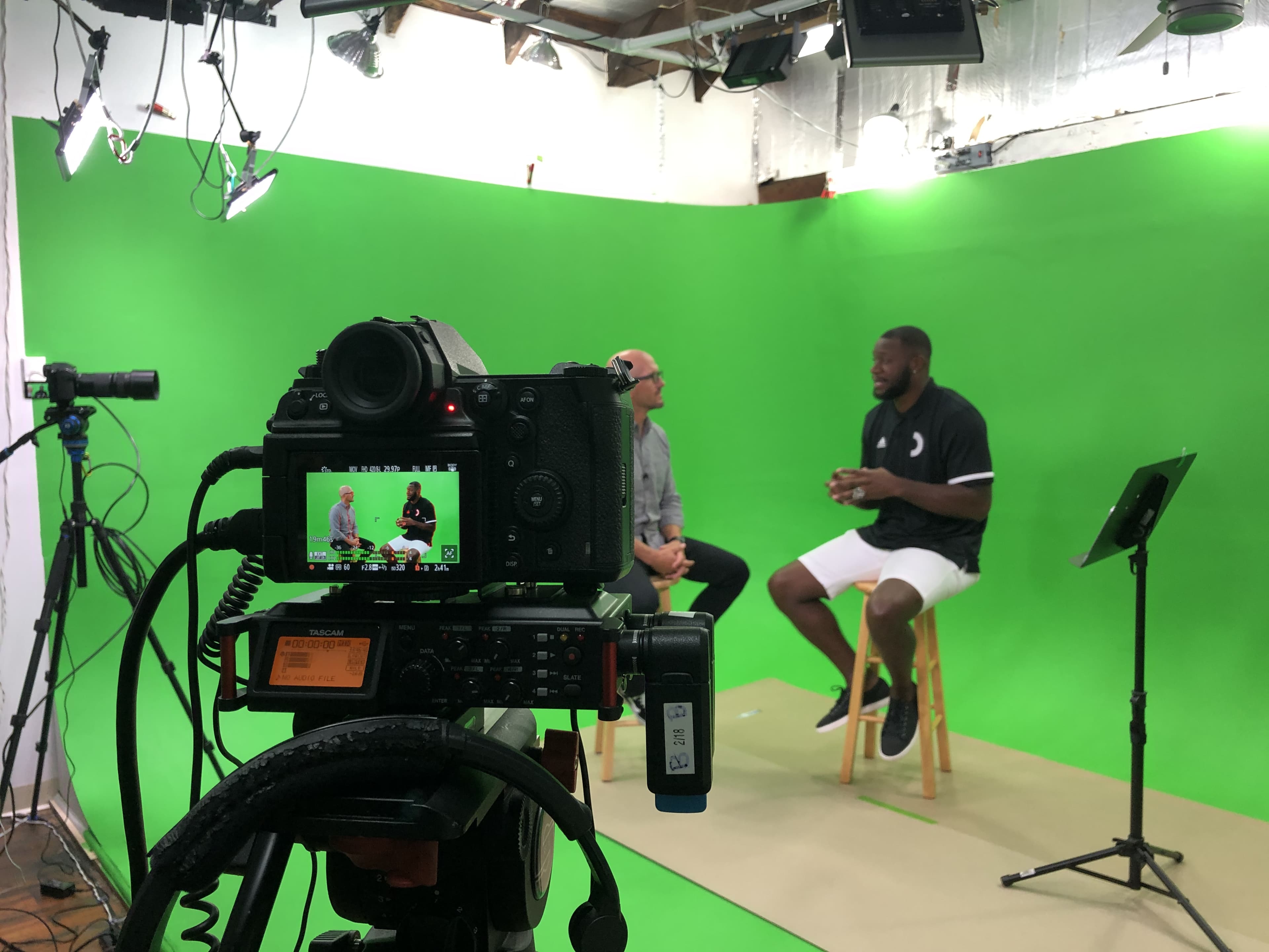 A camera captures two individuals seated on stools in front of a green screen, with studio lighting and equipment visible in the background.