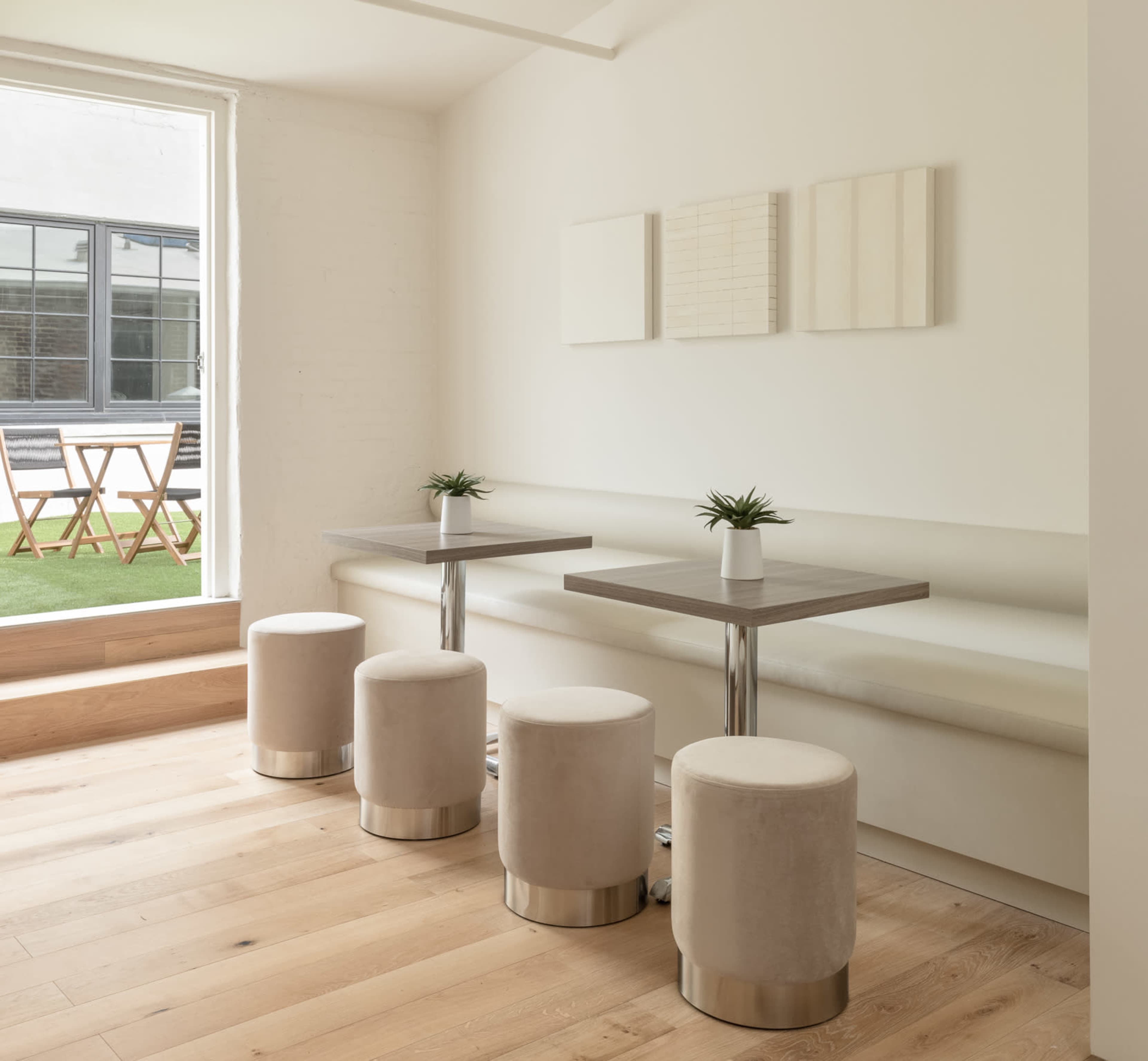 A bright, minimalist interior features a light wooden floor, a simple table with metal legs, and four upholstered stools arranged around it.