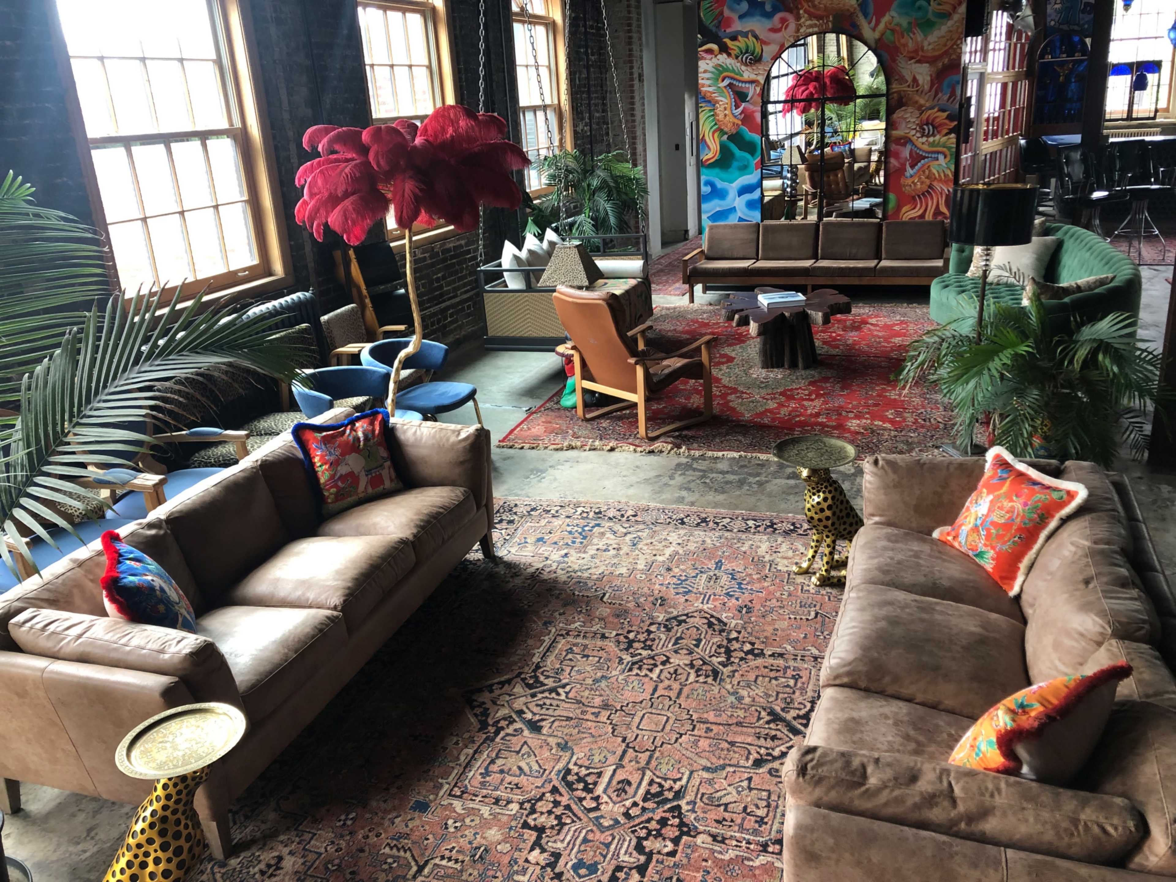 The image shows a stylish indoor lounge area with two brown sofas, colorful cushions, a large rug, and tropical plants, set against a backdrop of vibrant wall art.