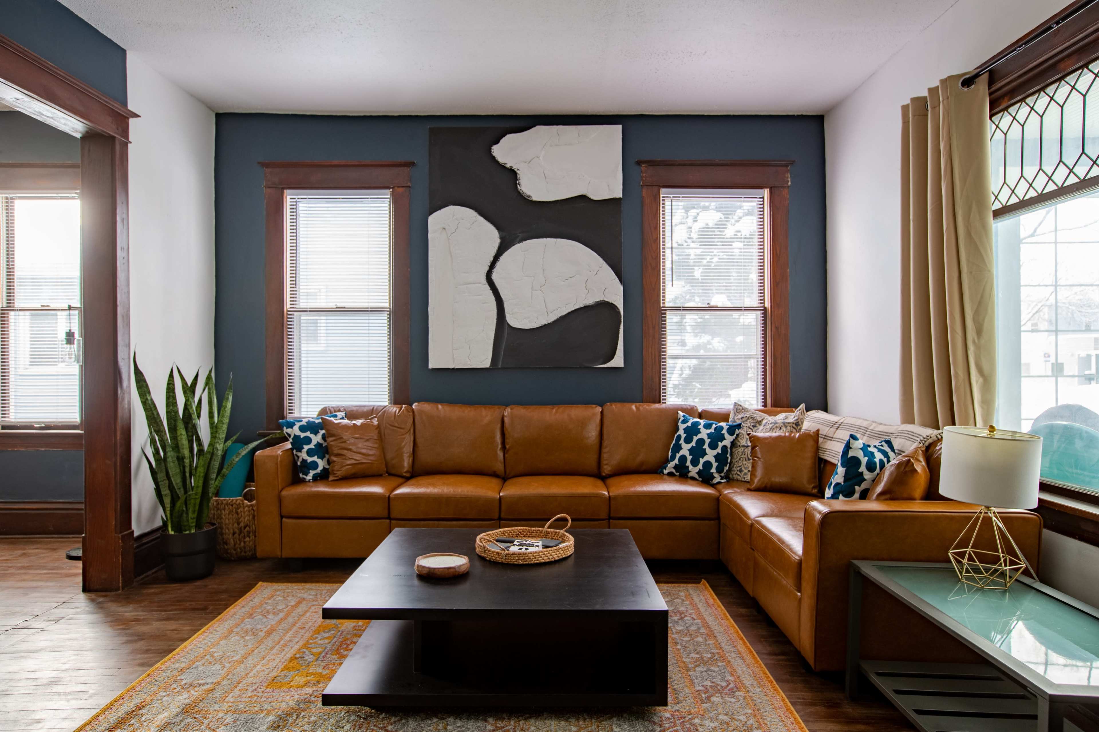 The image shows a cozy living room featuring a brown sectional sofa, a black coffee table, and a large abstract art piece on the wall, with plants and natural light coming from the windows.