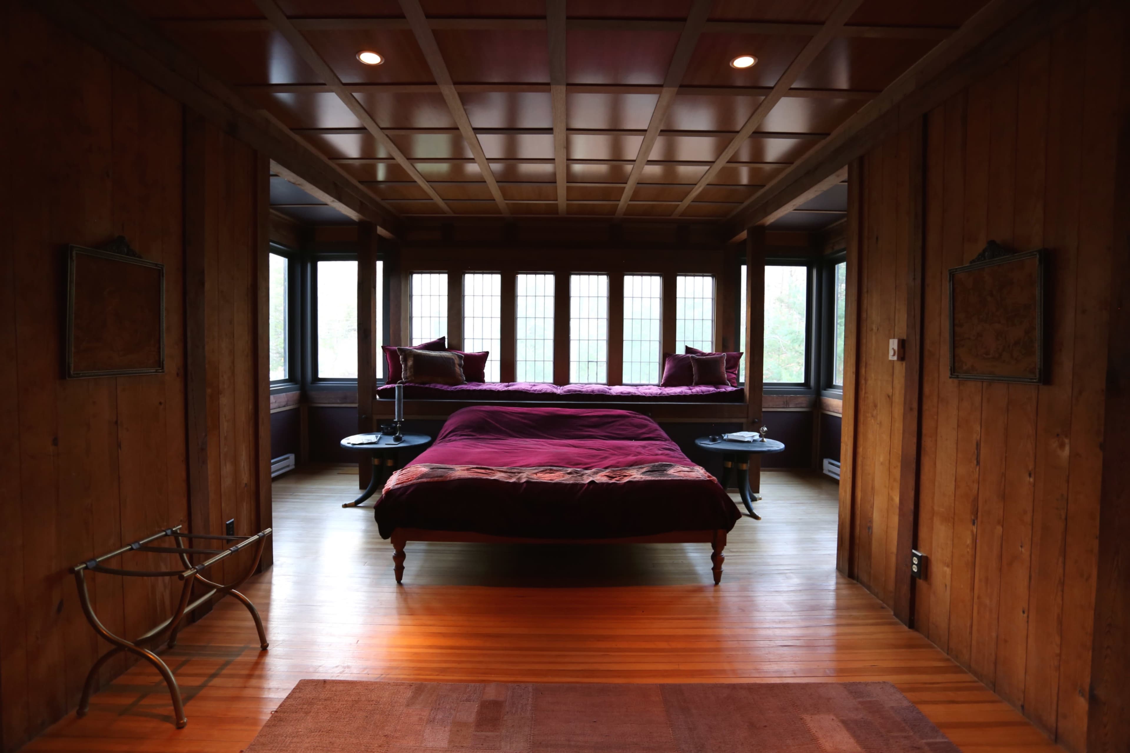 A spacious room features two beds with purple bedding, surrounded by wooden walls and large windows.