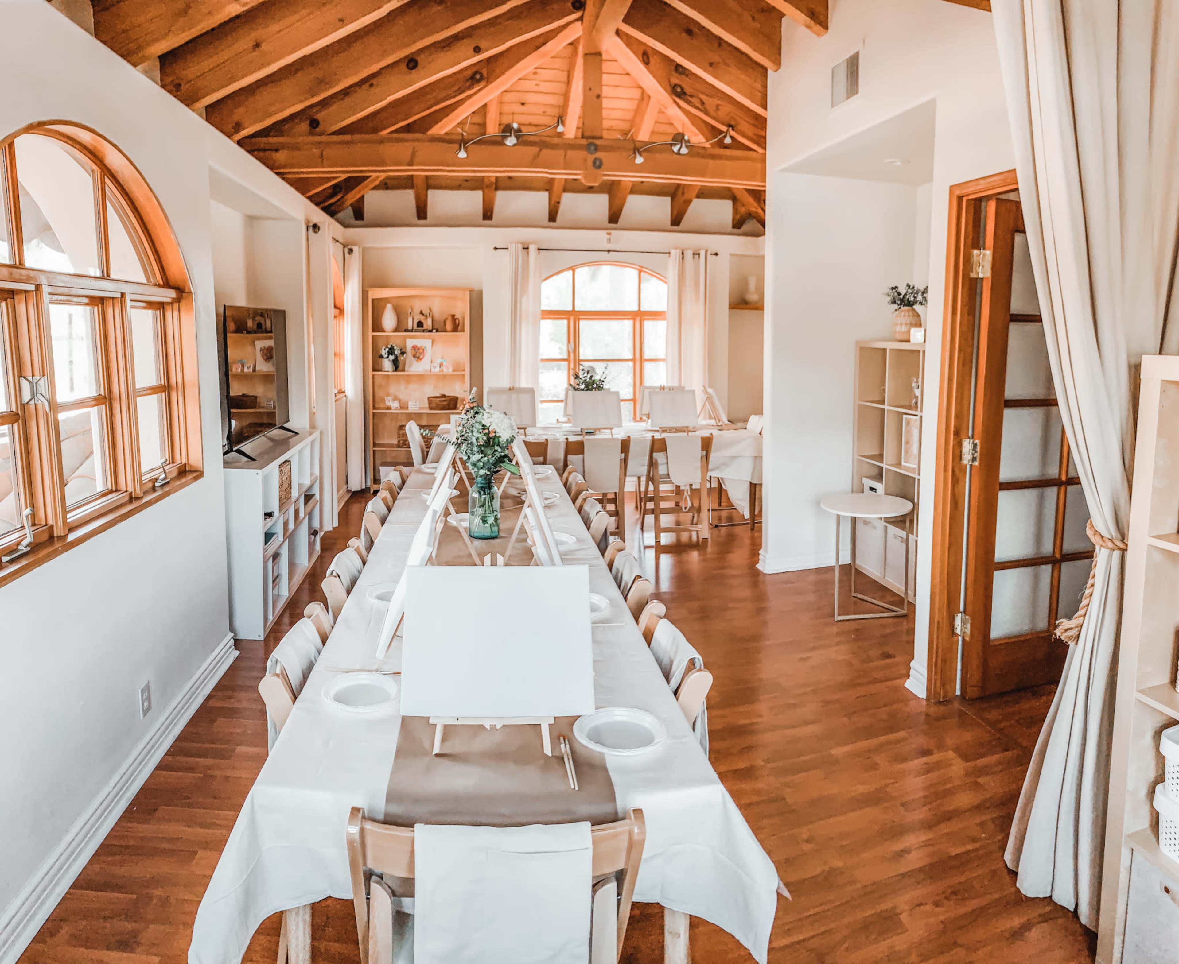 A spacious room with a long wooden table set for dining, surrounded by light-colored walls and accented by wooden beams in the ceiling.