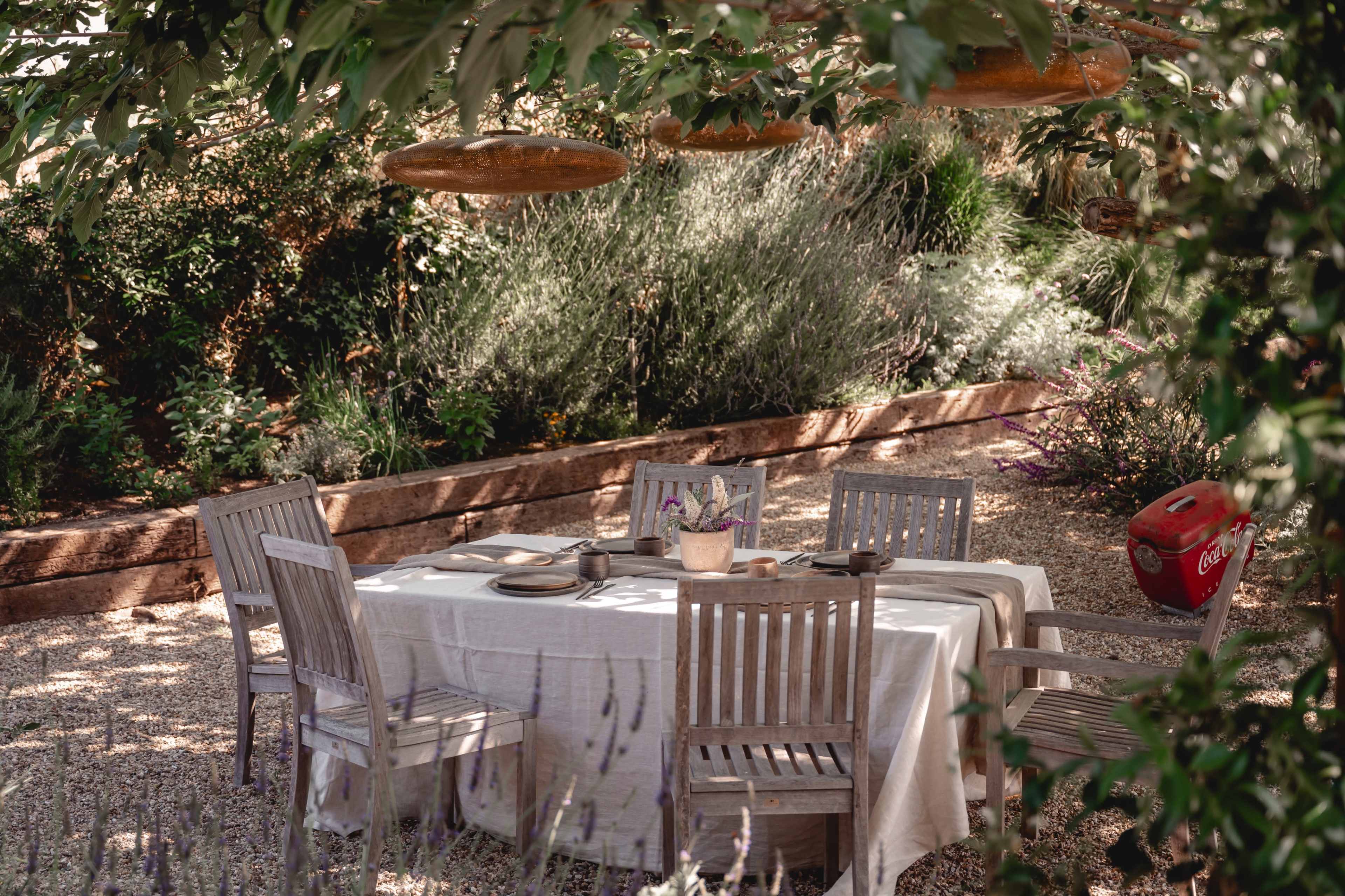 A wooden table is set with plates and utensils, surrounded by rustic chairs under greenery in a garden.