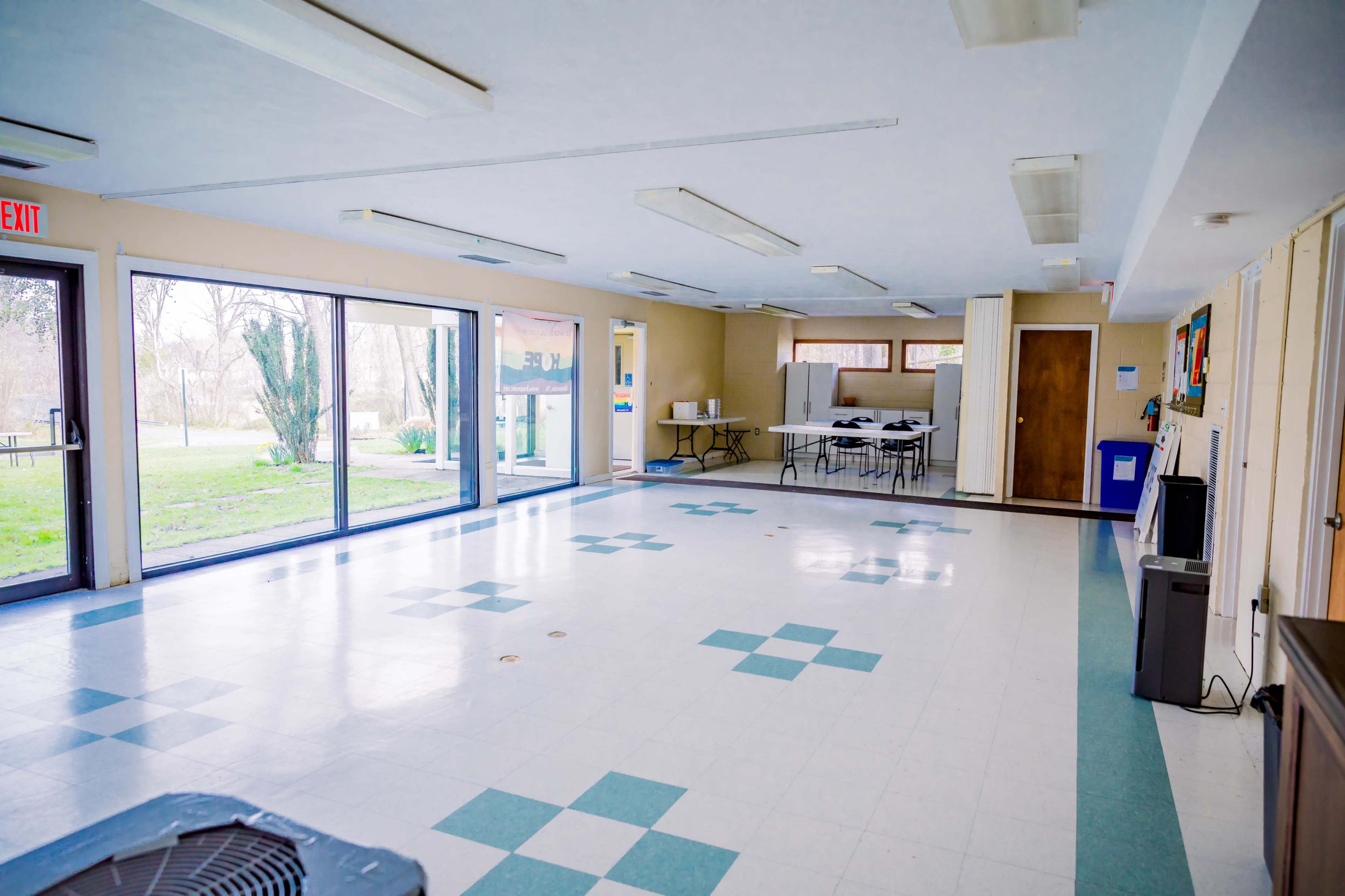 A spacious, empty room with tiled flooring, large windows, and tables set up against the wall.