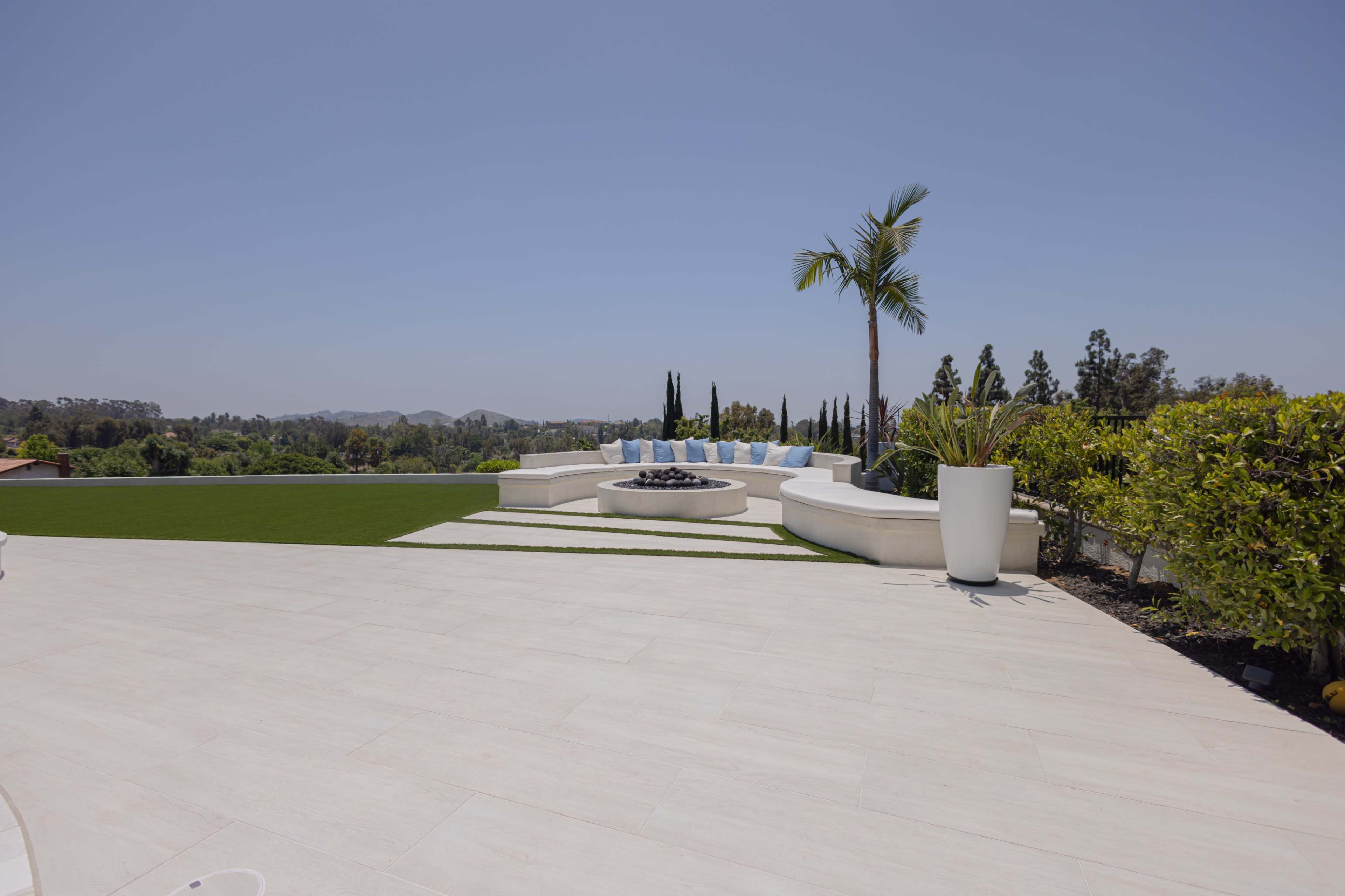 A spacious outdoor patio with white stone flooring, featuring a circular seating area around a fire pit and bordered by green grass and landscaped plants.