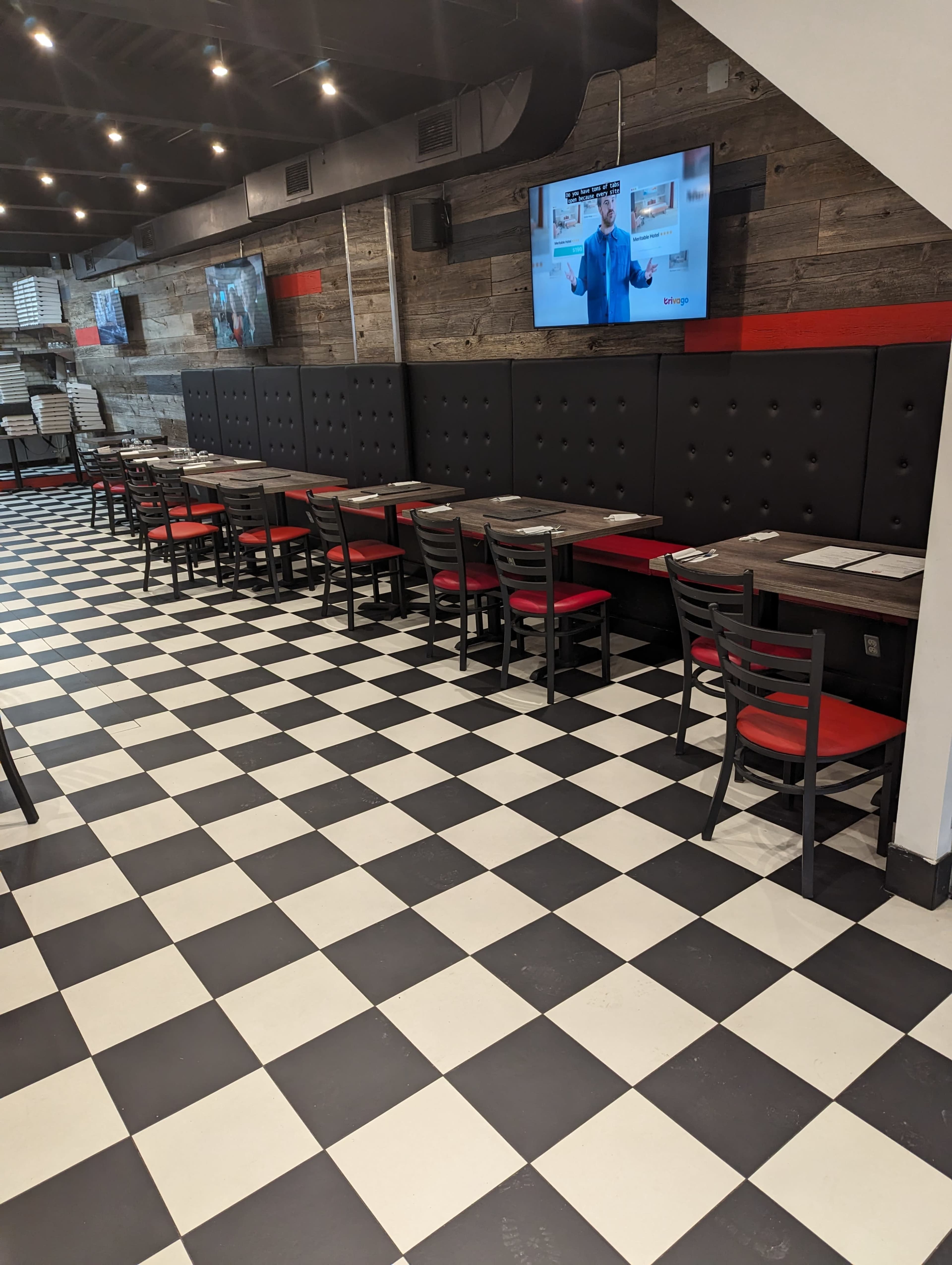 An empty dining area with black and white checkered flooring, red-accented booths, and two televisions mounted on the wall.