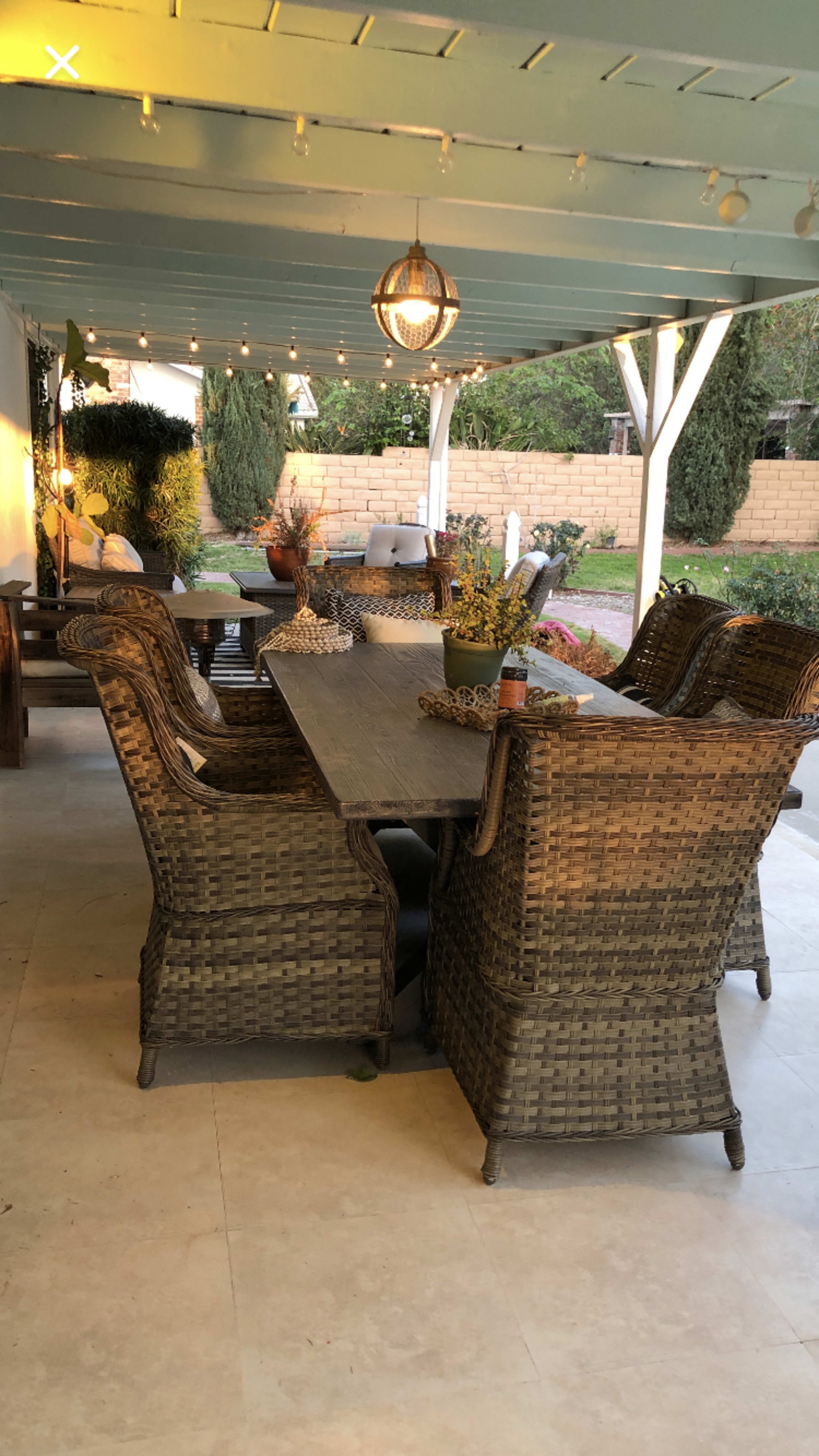 A covered outdoor dining area features a long table surrounded by wicker chairs, with string lights hanging above and a garden visible in the background.
