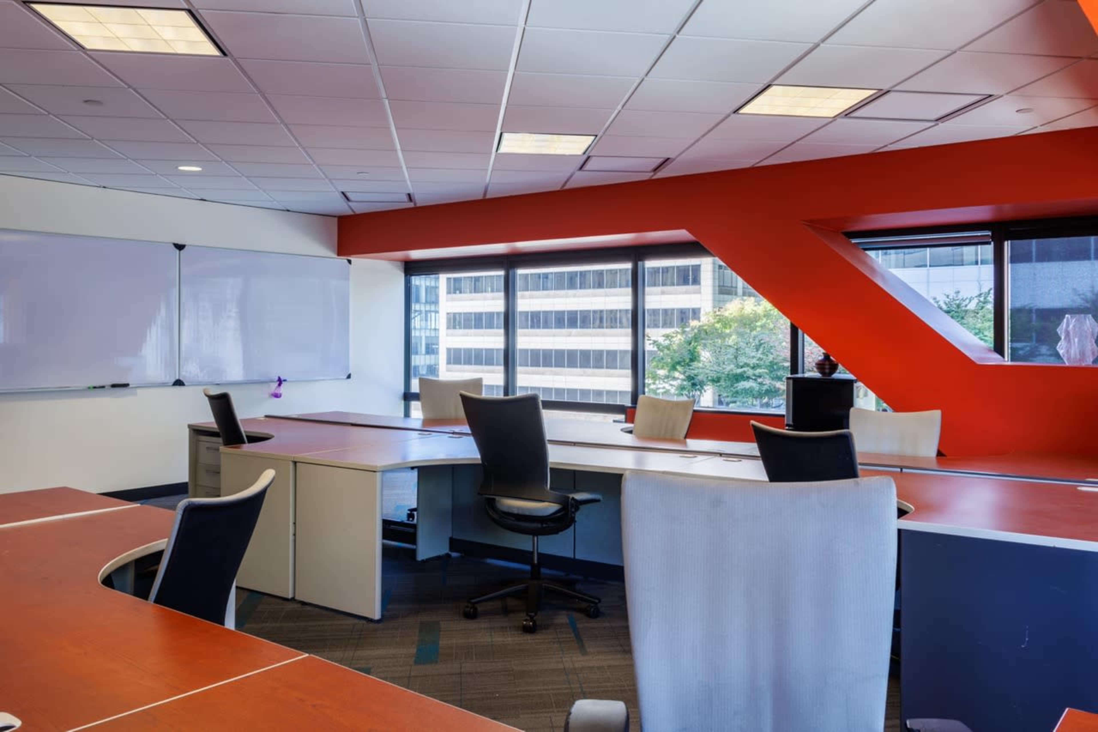 The image shows a modern office space with several desks and ergonomic chairs arranged around large windows, illuminated by natural light.