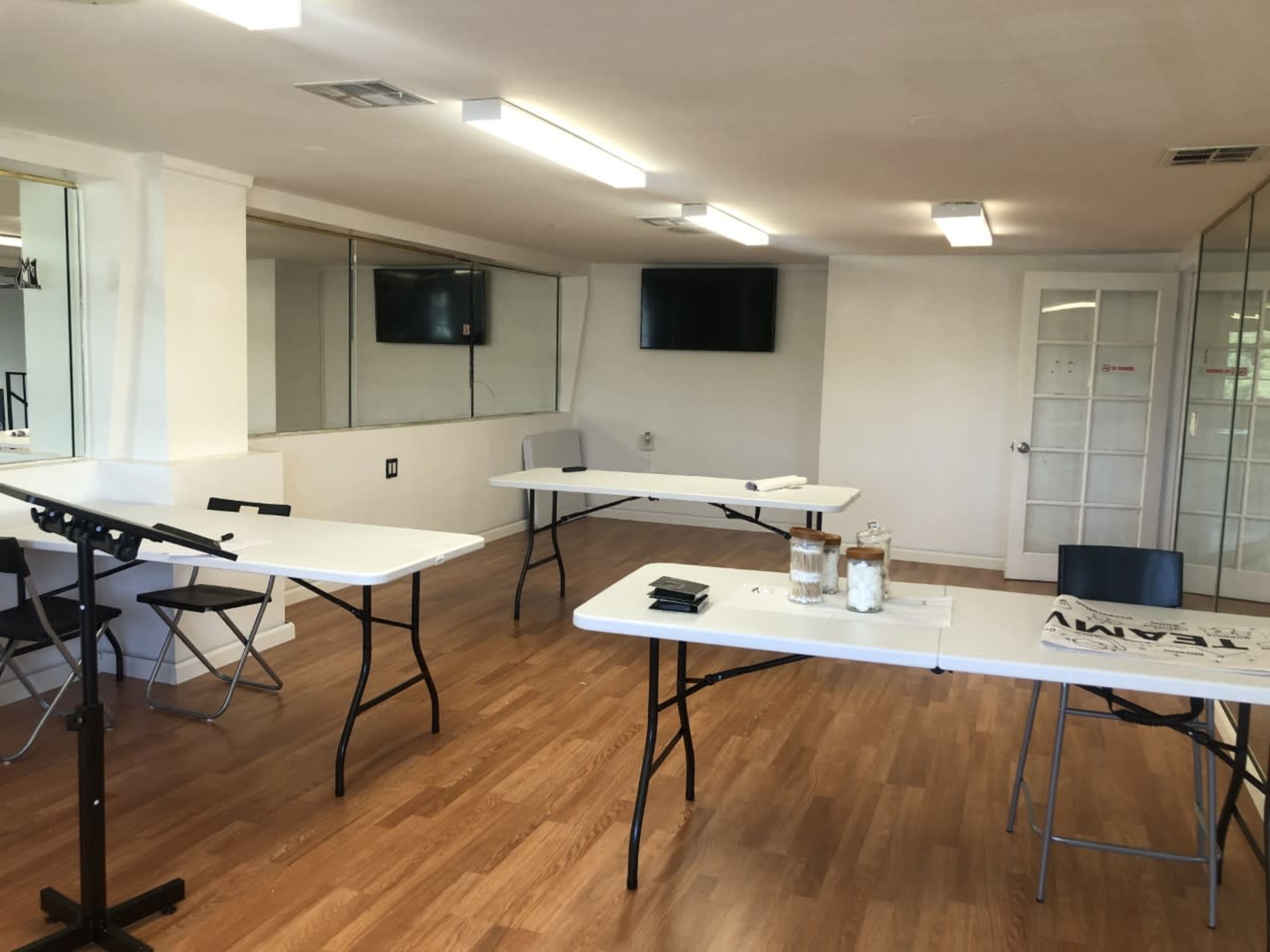 A room with wooden floors contains several folding tables and chairs, with two wall-mounted screens visible.
