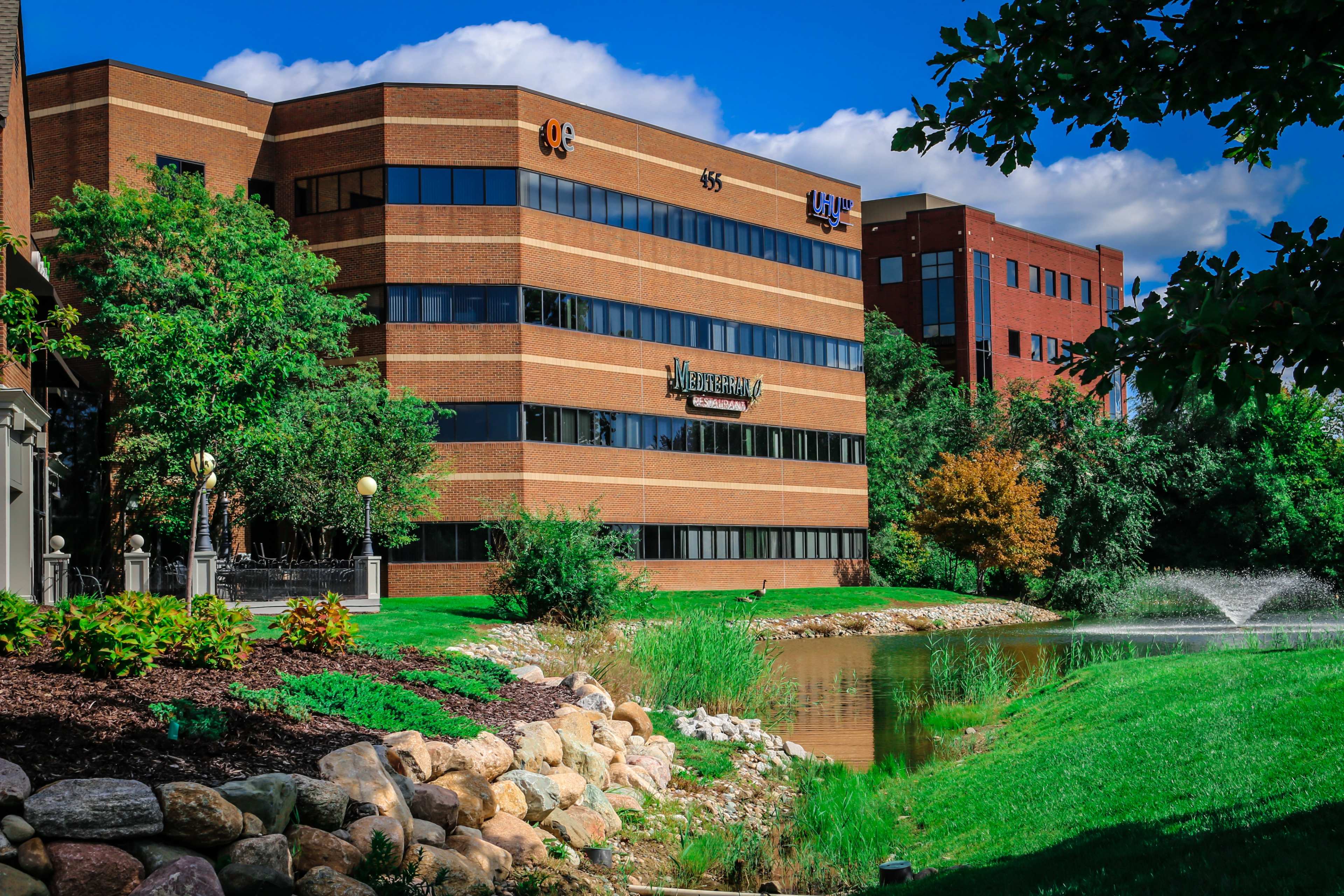 A brick office building alongside a pond, surrounded by greenery and trees with a blue sky in the background.