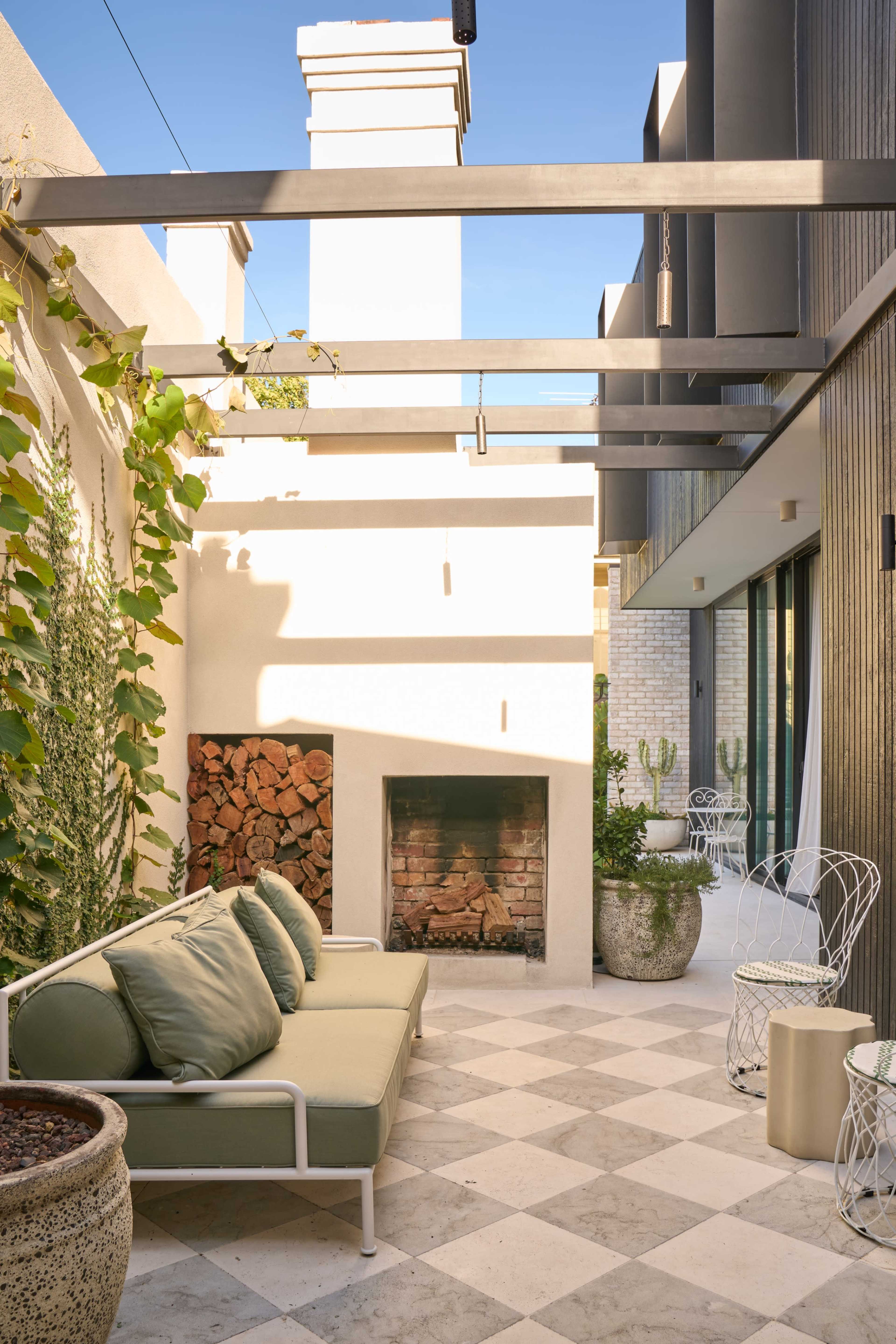 The image shows a patio area featuring a sofa with green cushions, a fire pit with stacked wood, and decorative planters, surrounded by modern architecture.