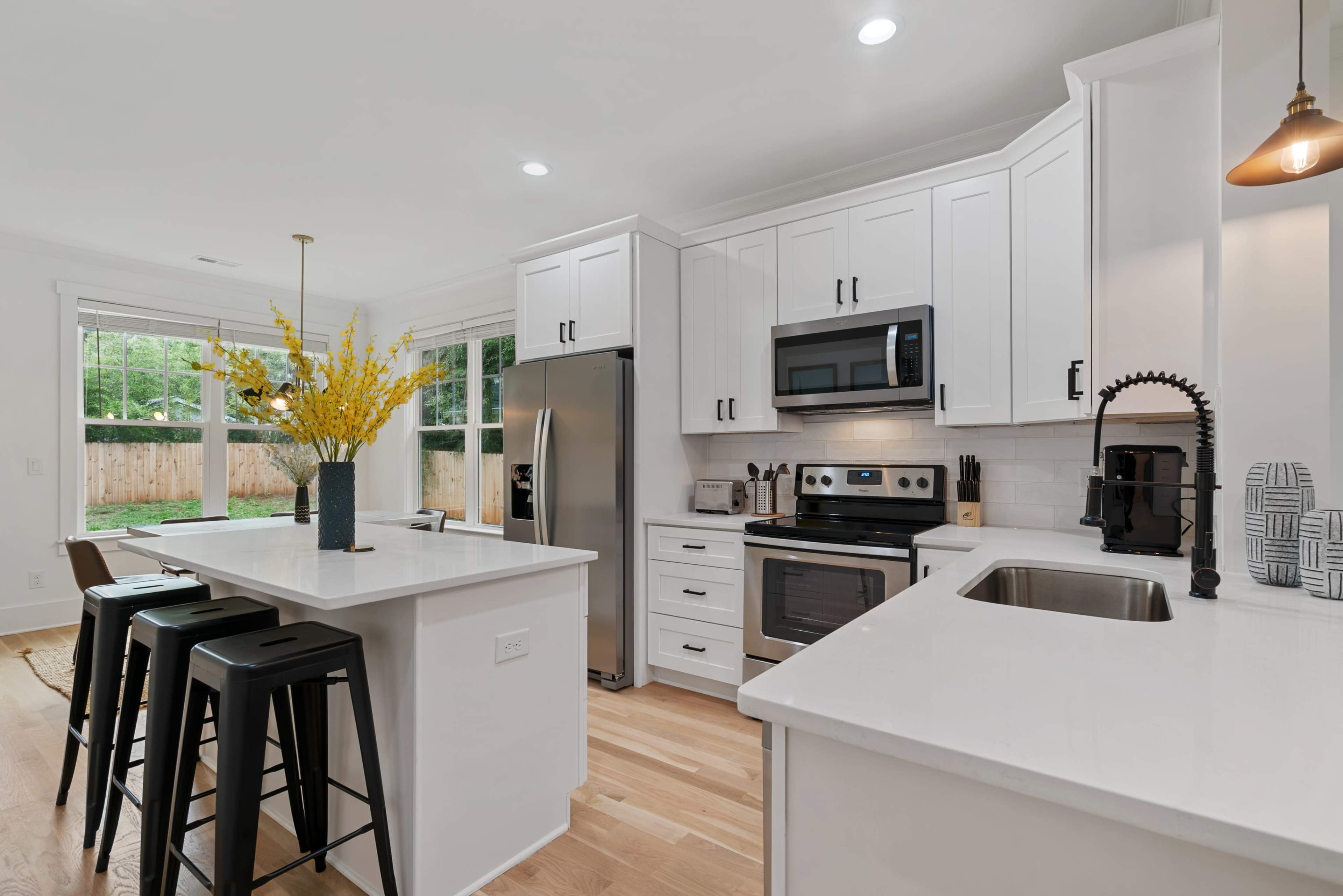 A modern kitchen with white cabinetry, stainless steel appliances, and a central island featuring black stools and a vase of yellow flowers.
