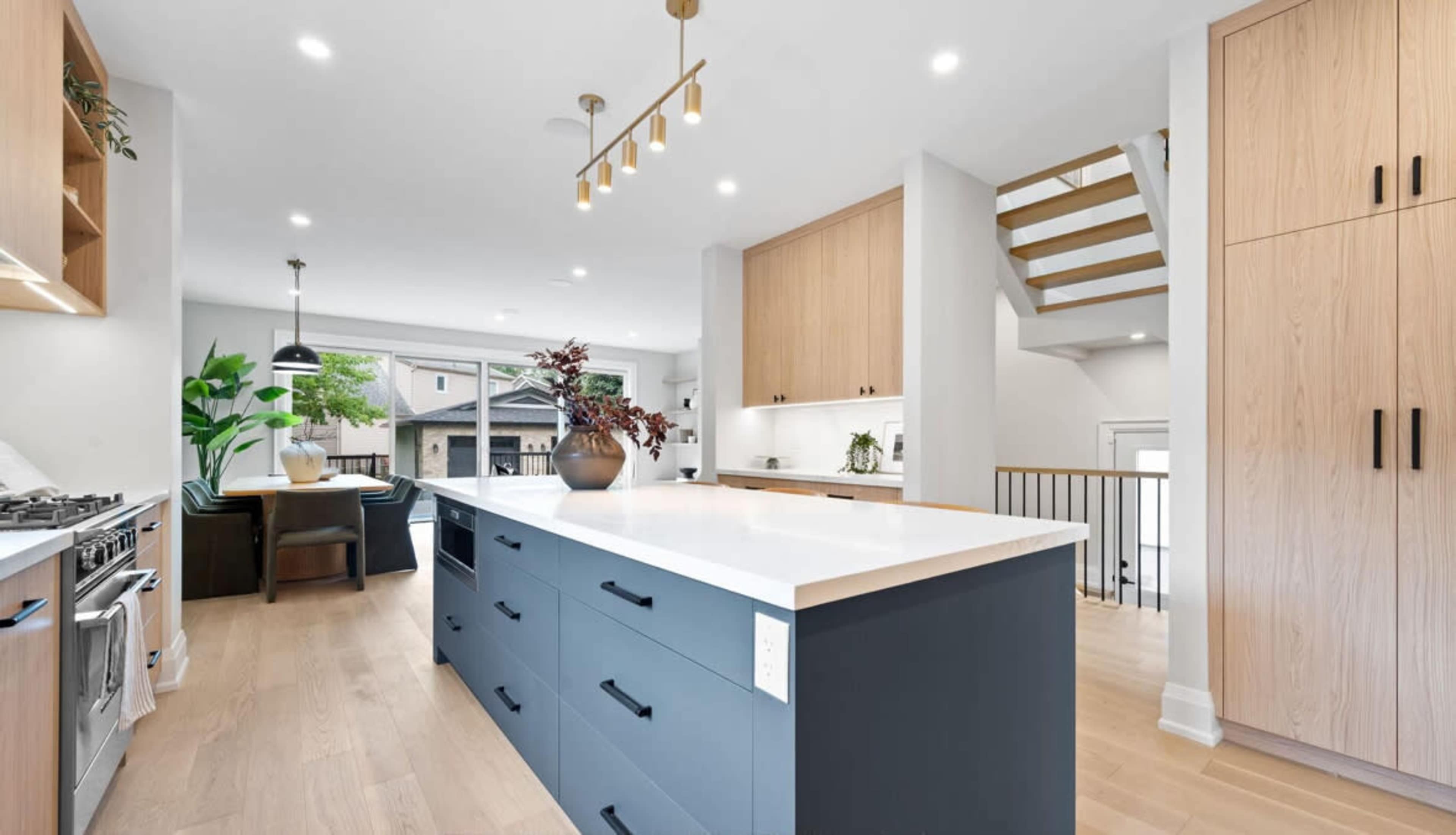 The image shows a modern kitchen with a large central island, light wood cabinetry, and a staircase in the background.