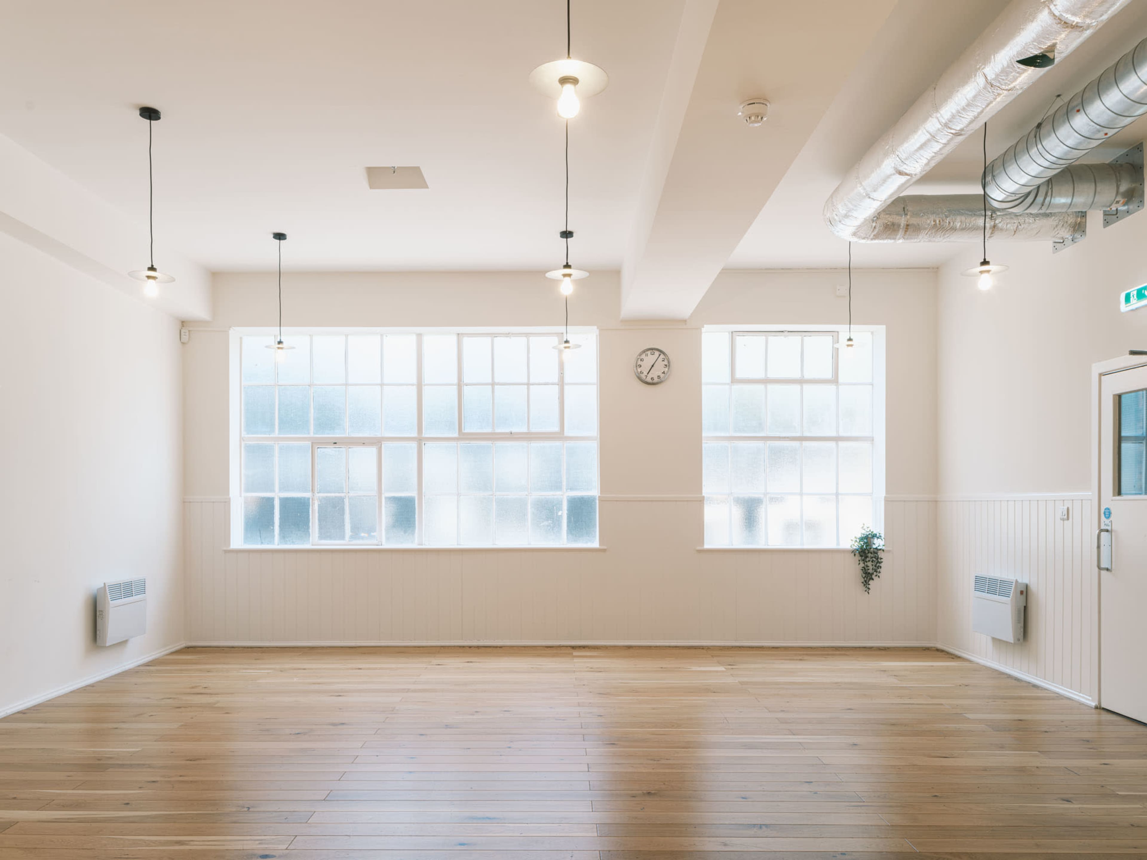 A spacious, empty room with wooden flooring, large windows, and industrial-style lighting fixtures.