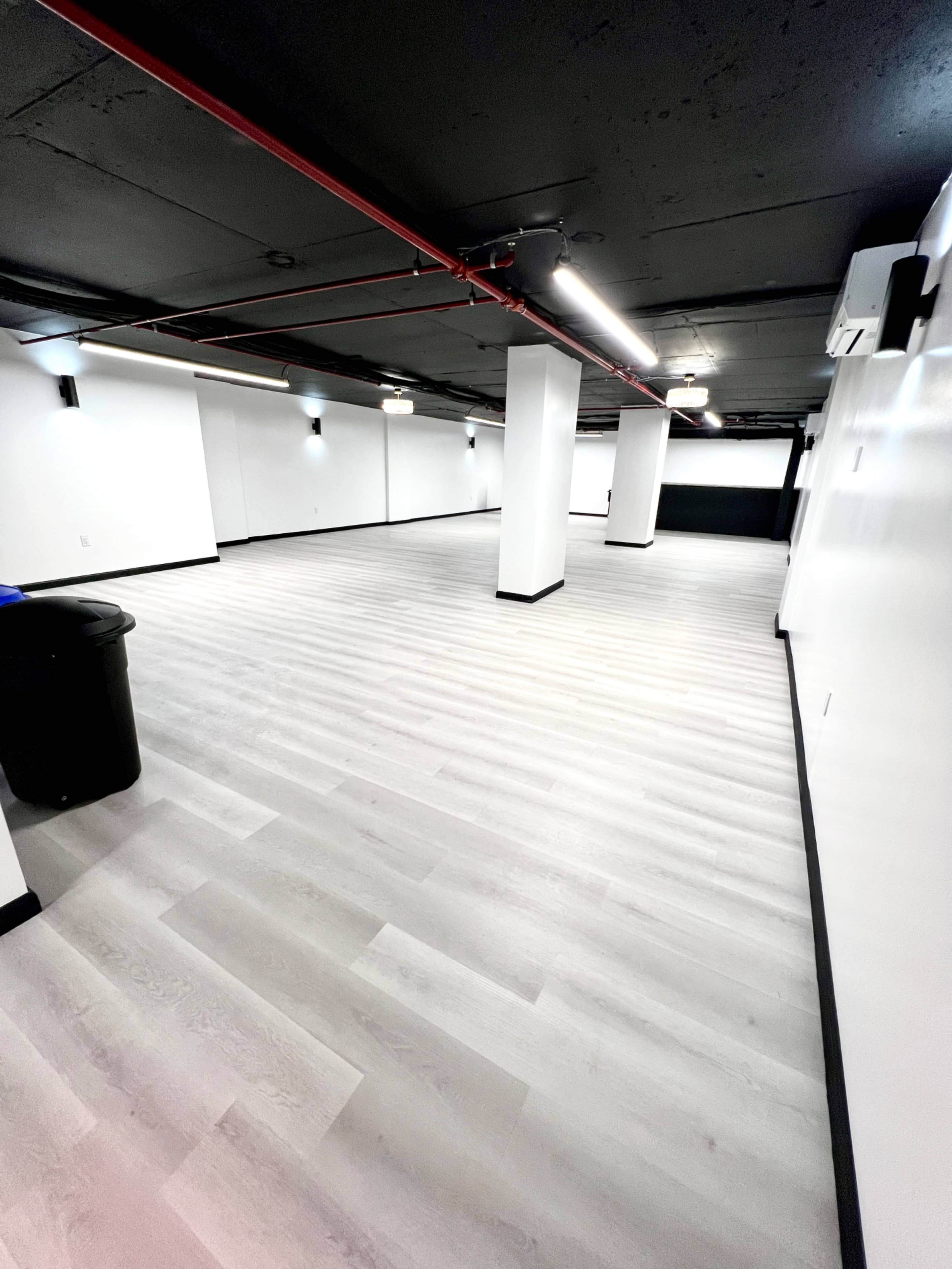 A large, empty room with gray wooden flooring and white walls, featuring exposed red pipes along the ceiling.