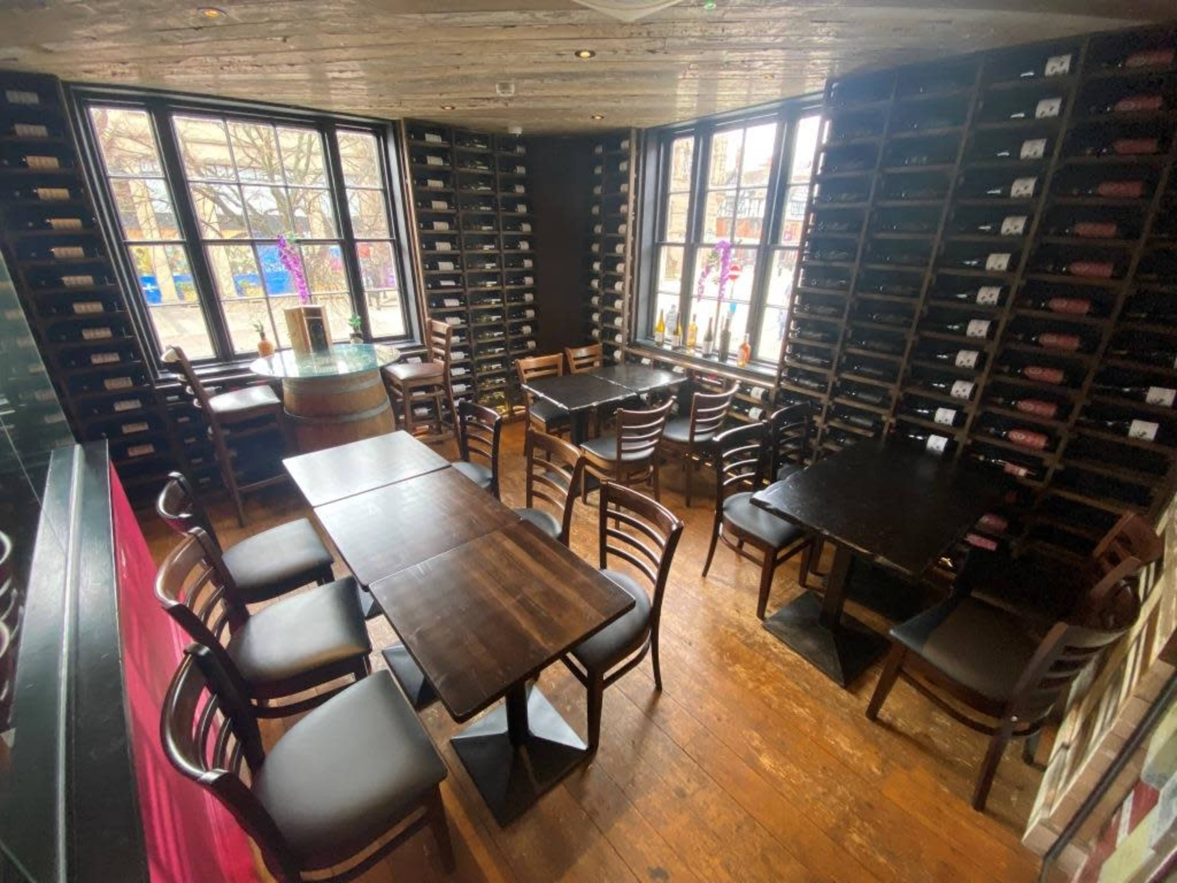 A restaurant interior with wooden tables and chairs arranged around a spacious layout, featuring large windows and wine racks lining the walls.