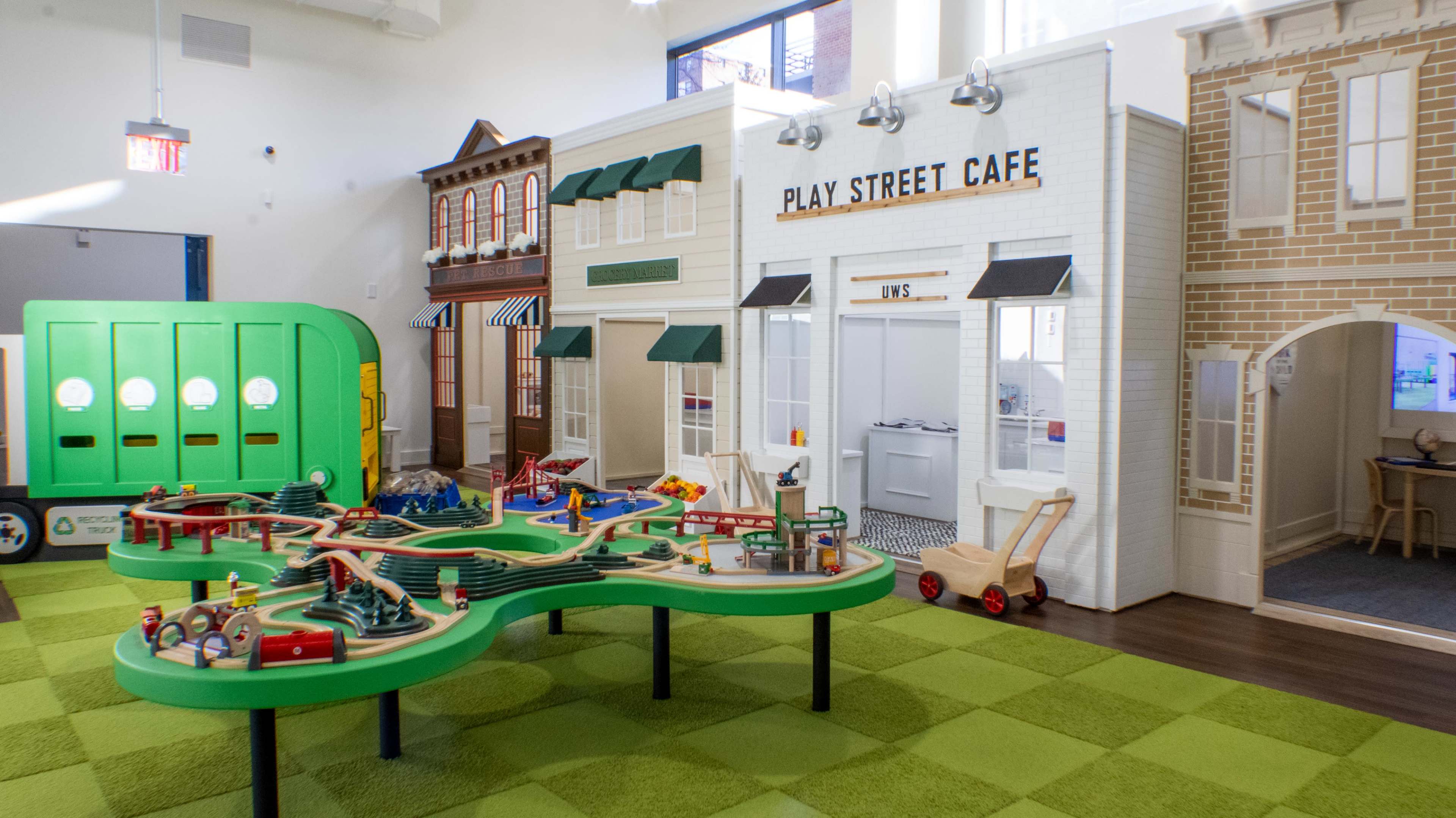 A brightly colored indoor play area designed for children, featuring toy buildings like a café and a train set on a green play mat.