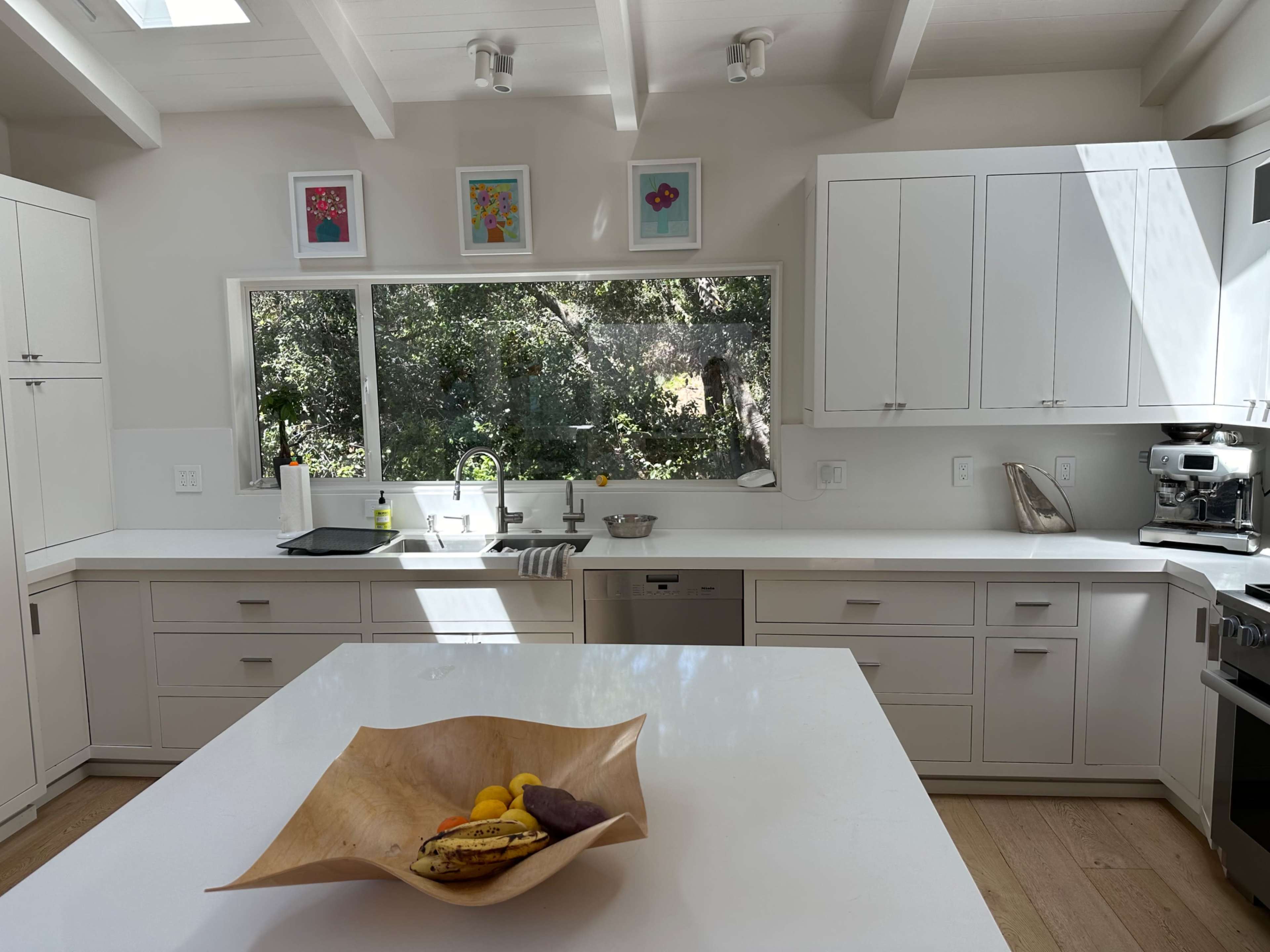 A modern kitchen features white cabinetry, a large island with a wooden bowl of fruit, and a view of greenery through a large window.