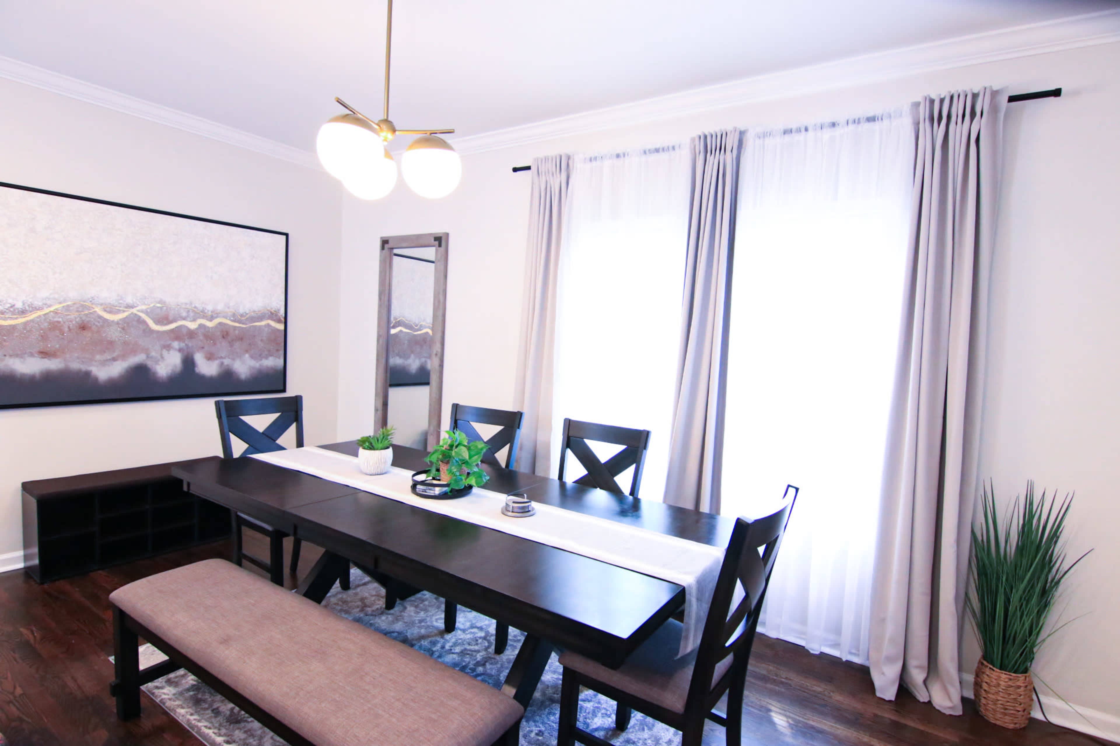A dining room features a long wooden table surrounded by x-back chairs, with a bench on one side, a large artwork on the wall, and large windows dressed with curtains.