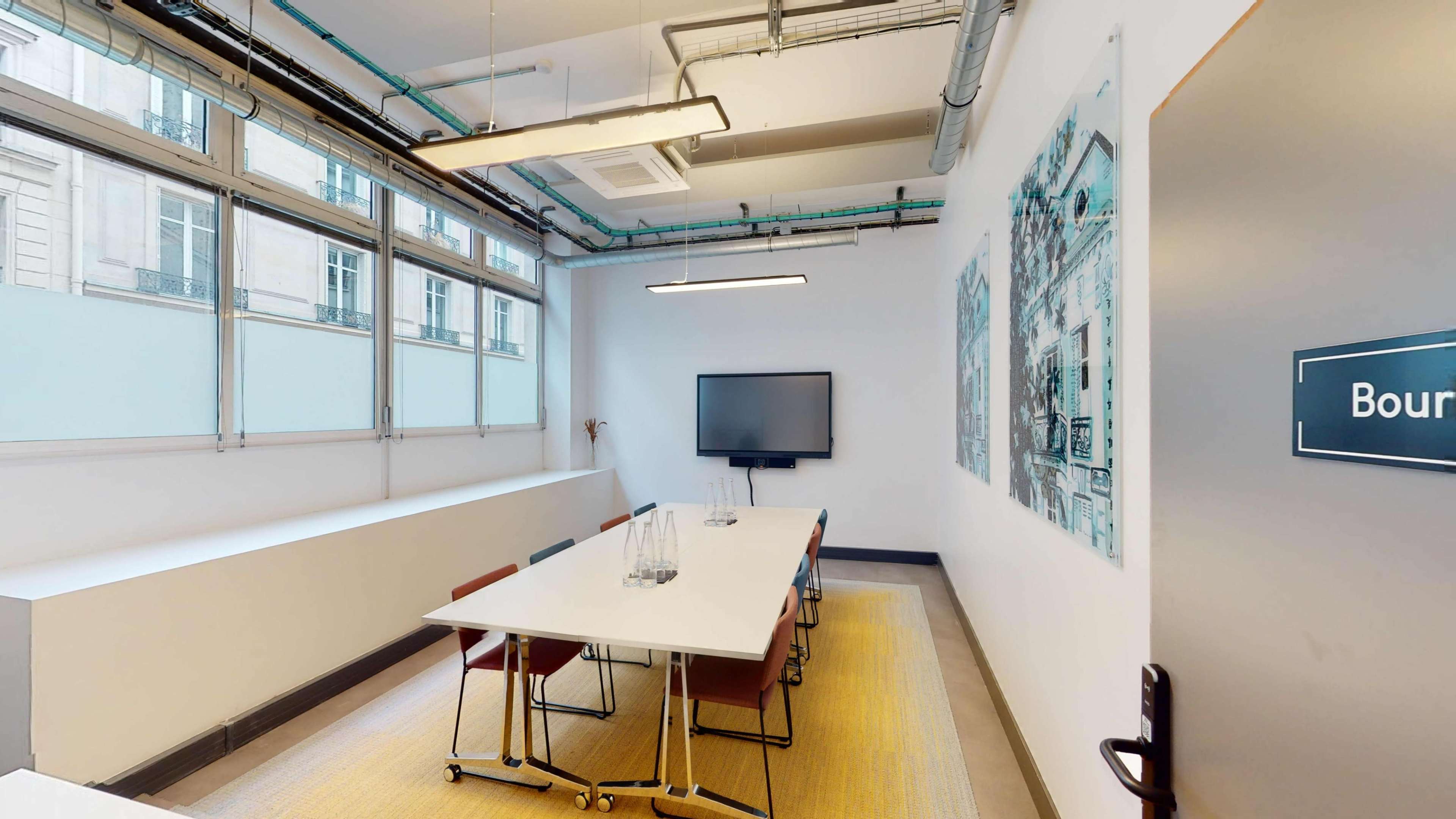 A meeting room features a long white table surrounded by chairs, a TV on the wall, and large windows allowing natural light.