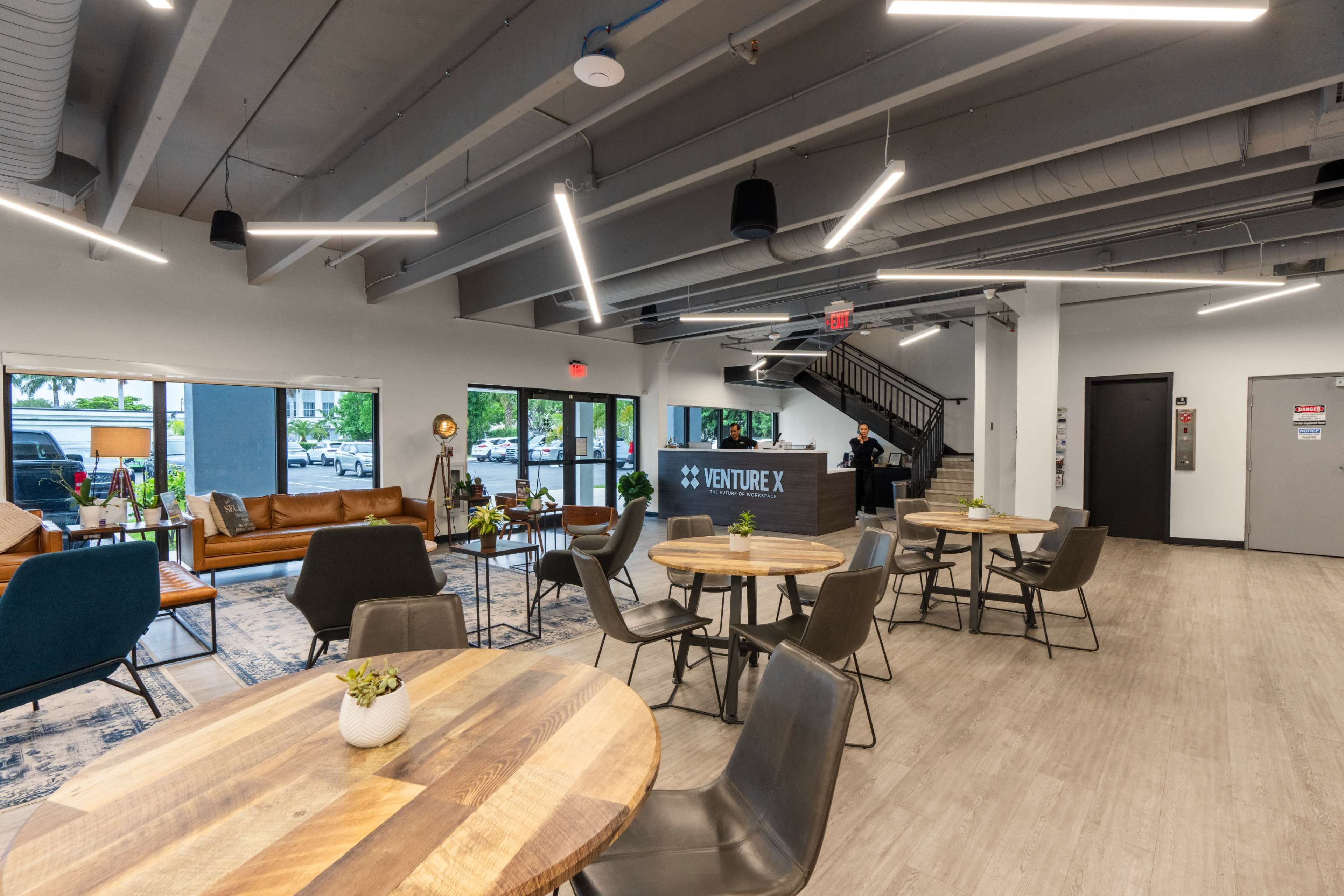 The image shows a modern office space with wooden tables, chairs, and a reception area marked "VENTURE X," featuring large windows that let in natural light.